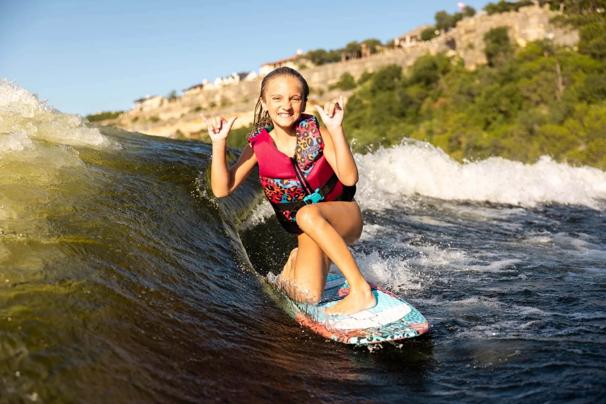 Smiling girl in a life jacket surfs a wave on a lake, making a rock on hand gesture, with trees and cliffs behind her.