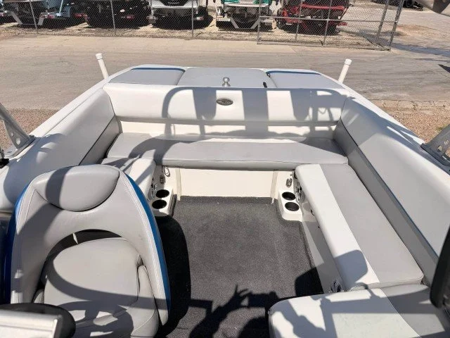 Interior of a boat with white and gray seating, a steering wheel, cup holders, and a gray carpeted floor, docked at a marina with cars visible in the background.