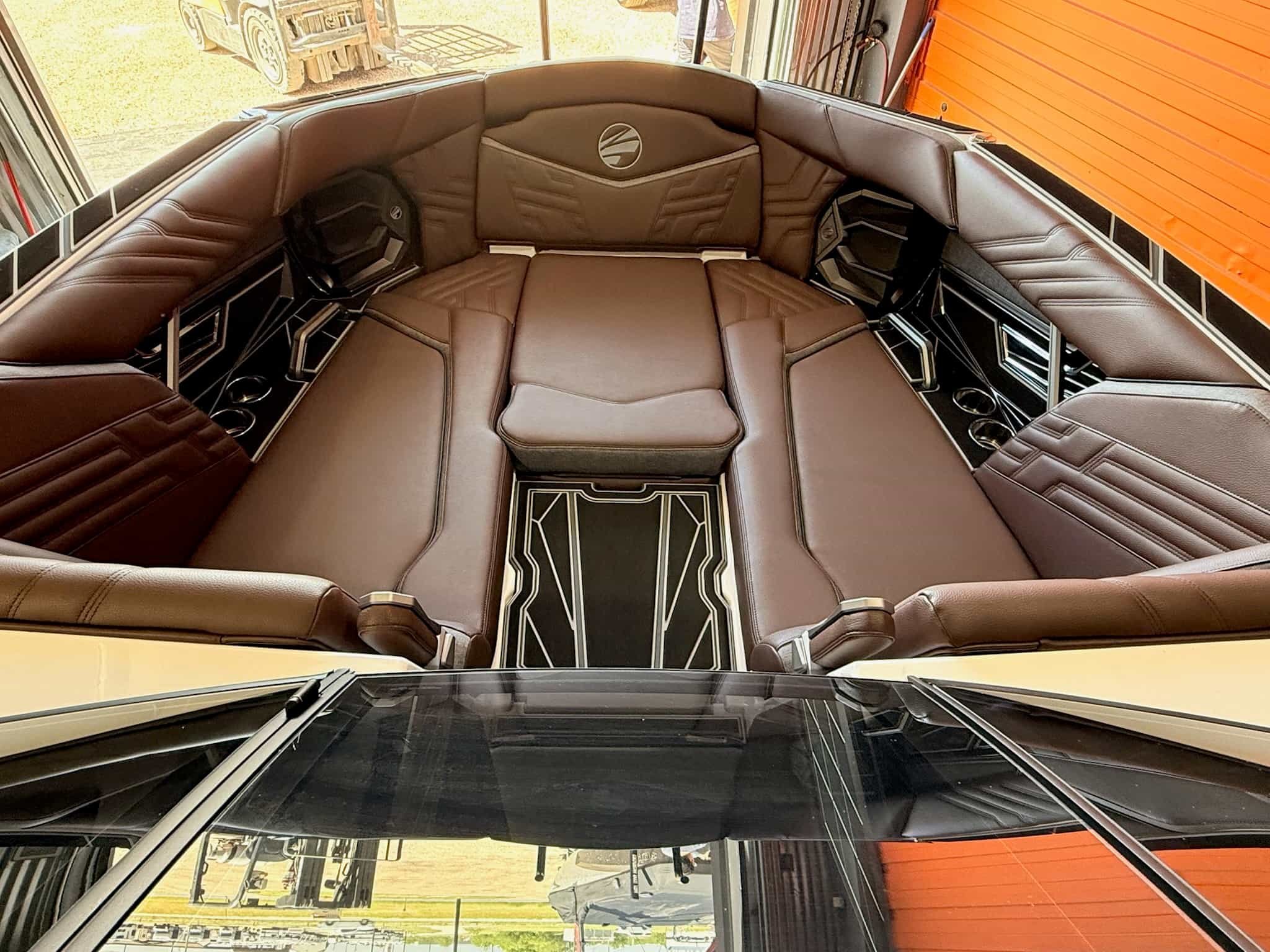 View from the front windshield of a black vehicle, looking at the interior of a brown leather sectional sofa with built-in speakers and a decorative logo on the backrest, inside a garage with a wooden wall and a sliding glass door.