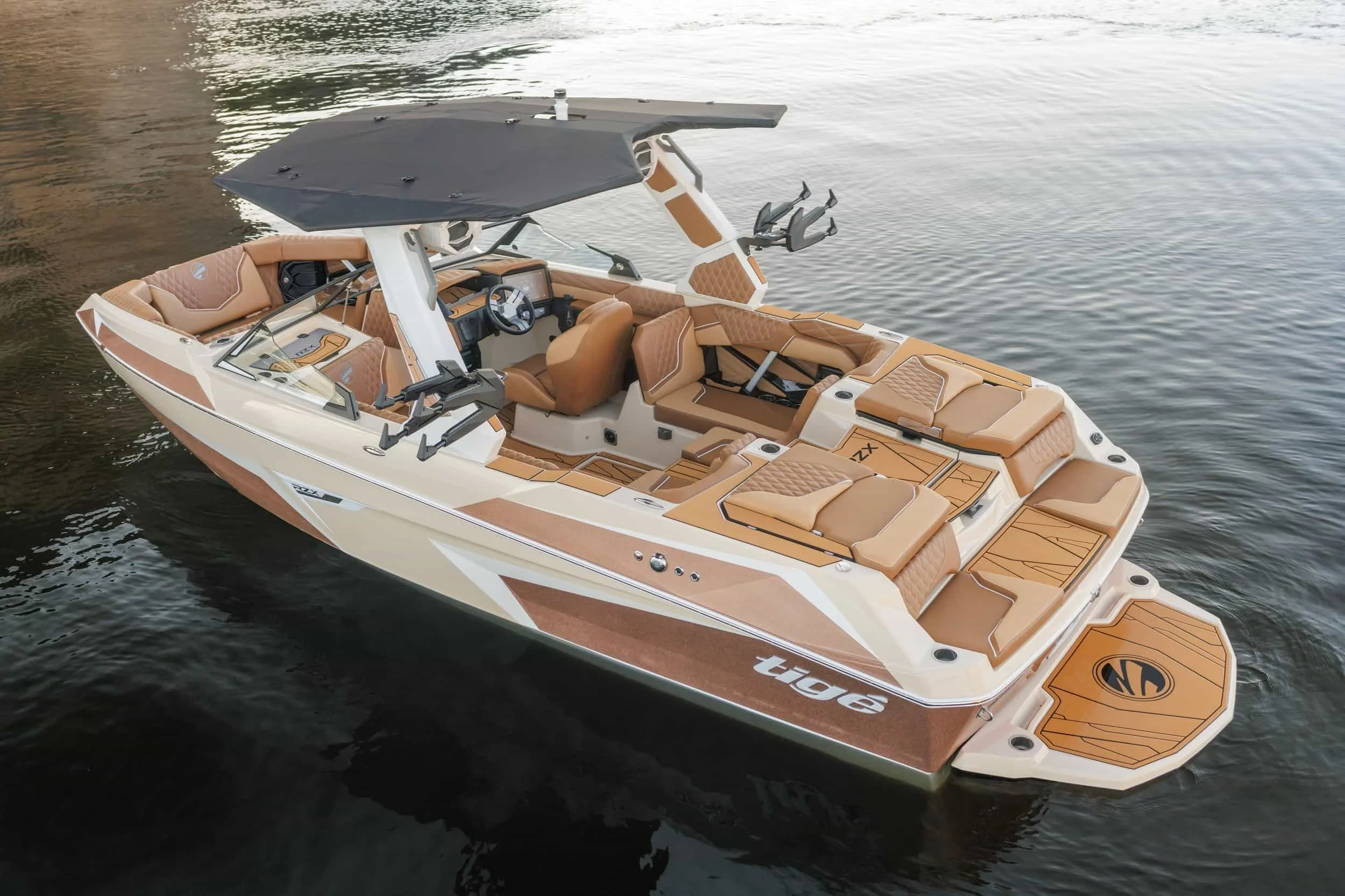 A luxury tan and white wake boat with cushioned seating and a black sunshade floats on calm water.