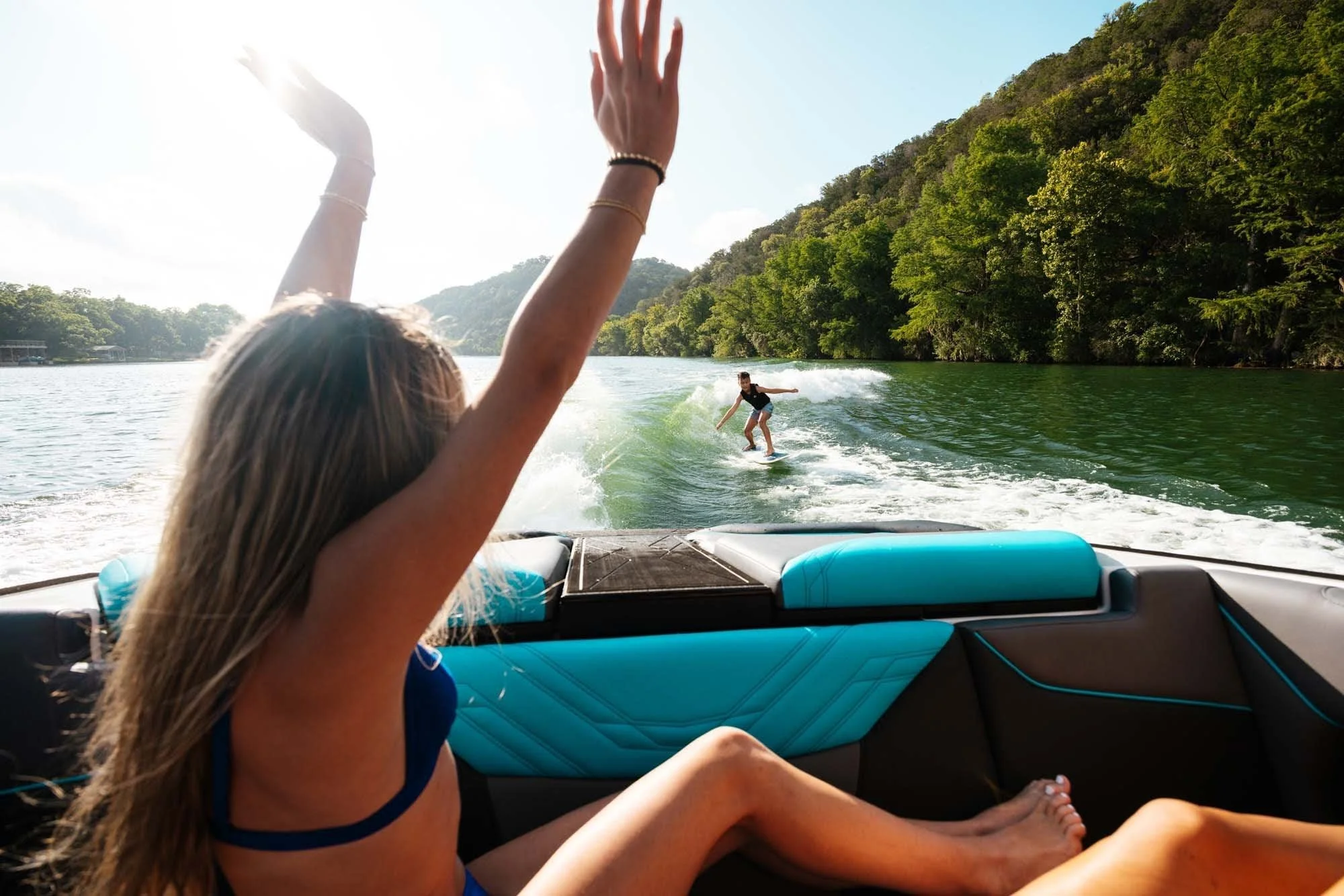 Person in a boat with arms raised, watching someone wakeboarding on a lake surrounded by green trees and hills under a sunny sky.