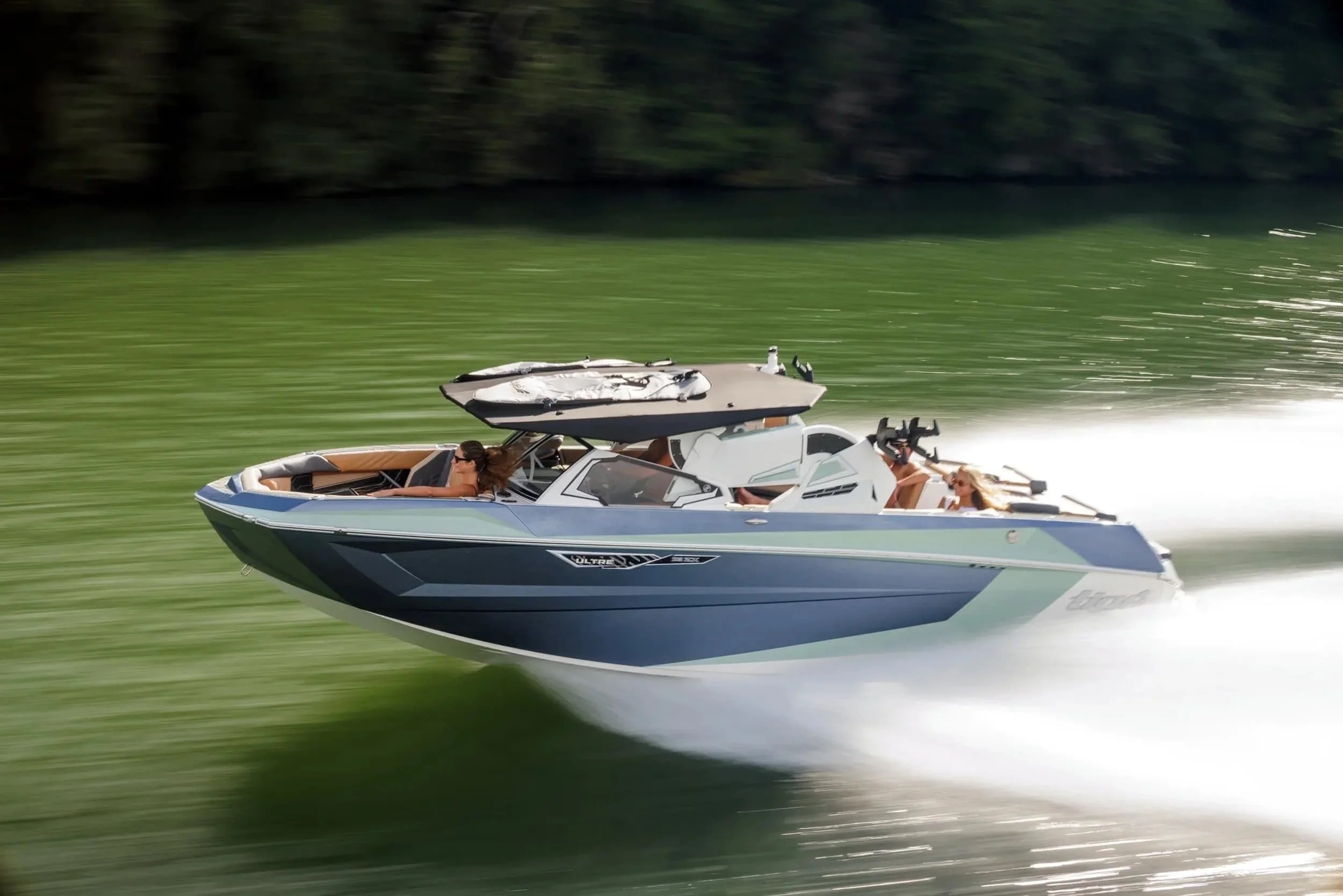 A blue and white speedboat moves quickly across green water, leaving a white wake behind. Trees line the background.