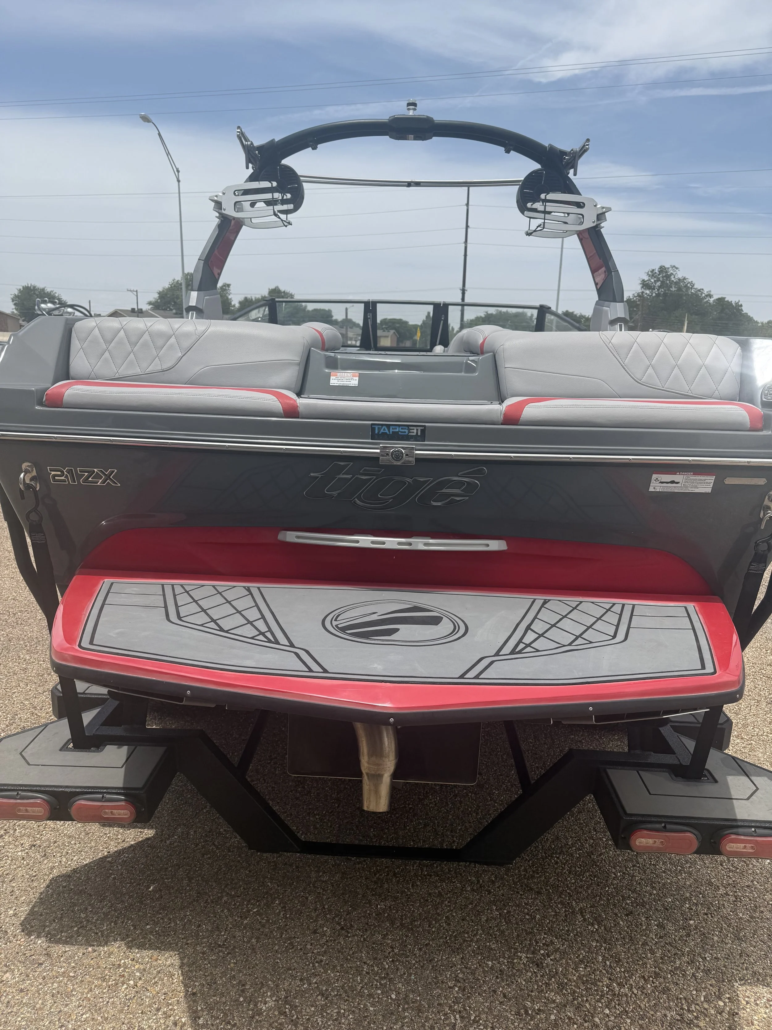 Front view of a boat with a gray and red color scheme, featuring a seating area with cushioned seats, a black TAPS-ET transducer, and a wakeboard tower with speakers and hardware. The boat is on a trailer on an asphalt surface, with power lines and t