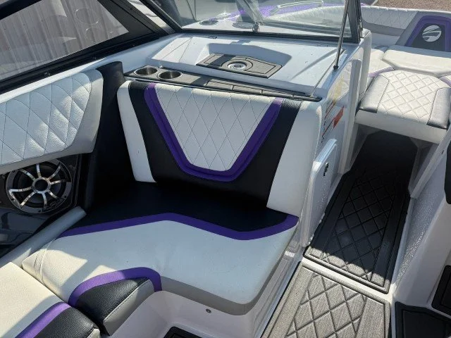 Interior of a boat with white and purple cushioned seating, diamond-stitch upholstery, and a control panel with a steering wheel, gauges, and controls.