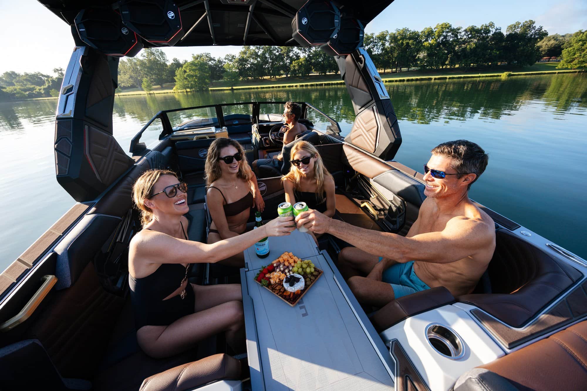 Four people in swimsuits toast drinks on a boat, enjoying snacks on a sunny day by a calm lake with trees in the background.