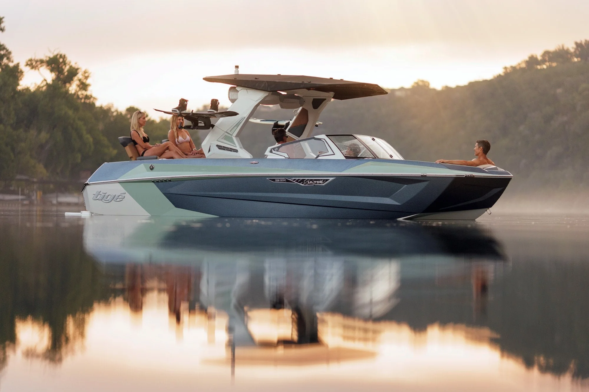 A sleek motorboat with people relaxing on board floats on calm water at sunset, surrounded by trees and soft reflections.