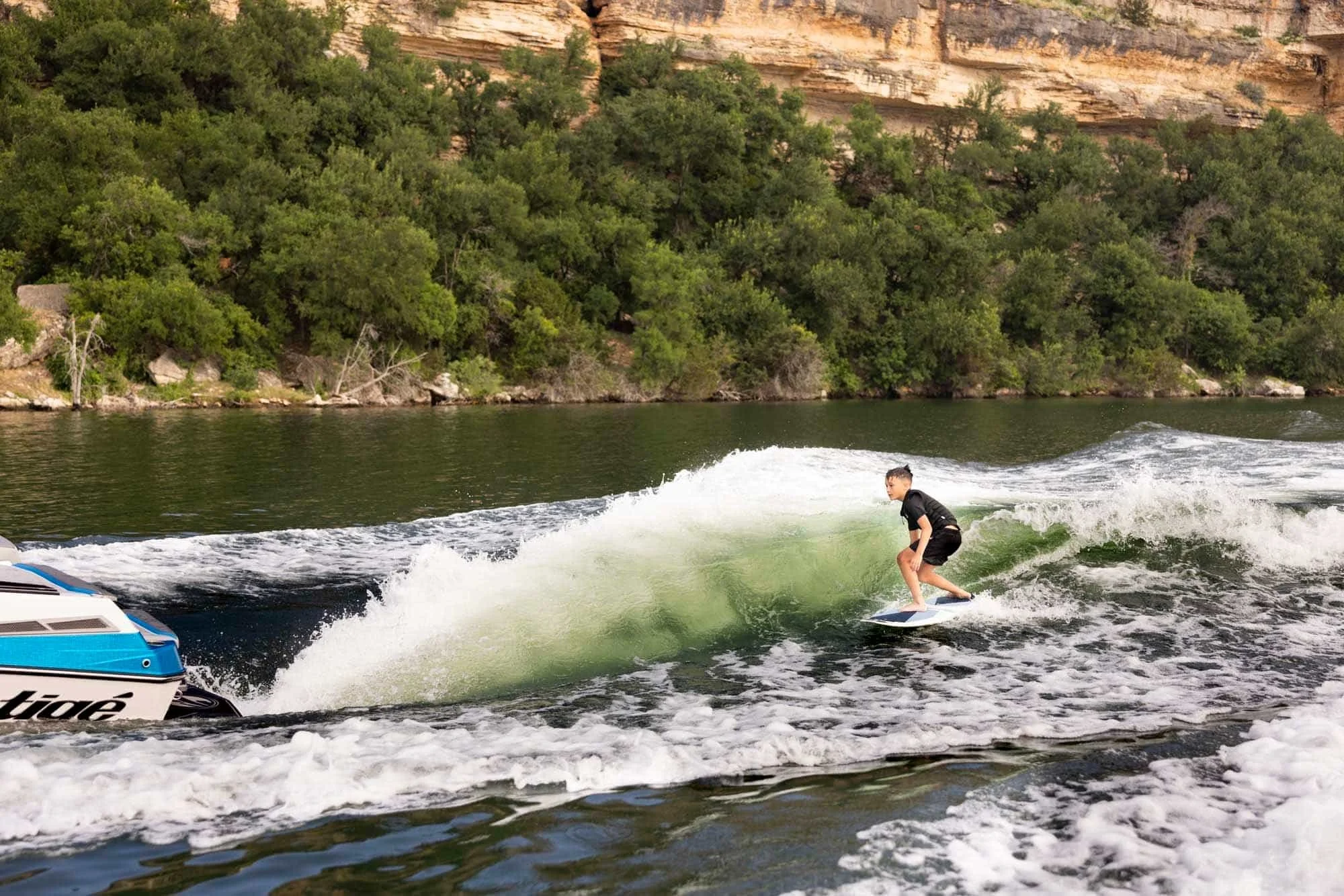 Man wakesurfs behind a boat on a river with green trees and rocky cliffs in the background.