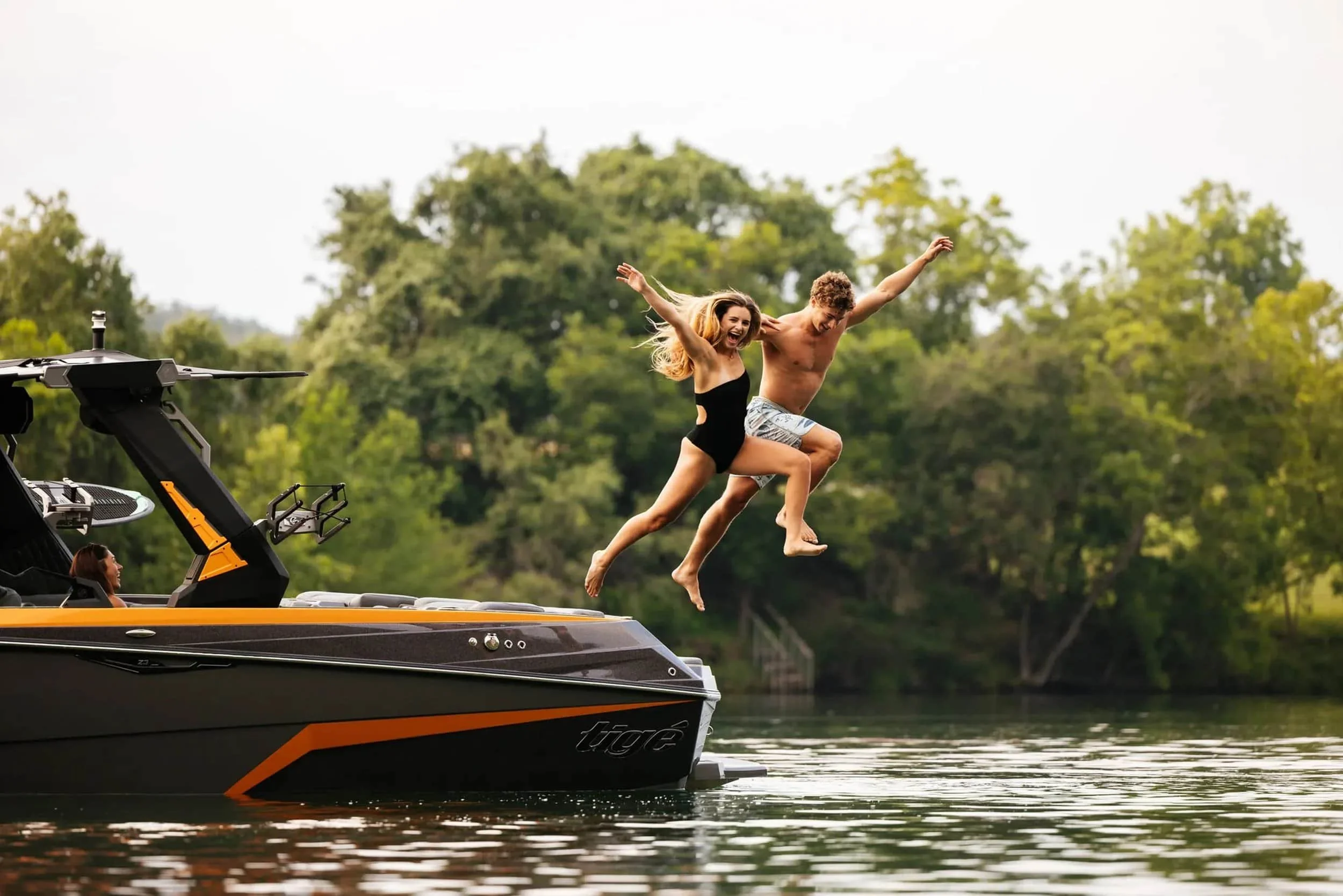 A man and woman in swimsuits joyfully jump off a boat into a lake, surrounded by trees on a sunny day.