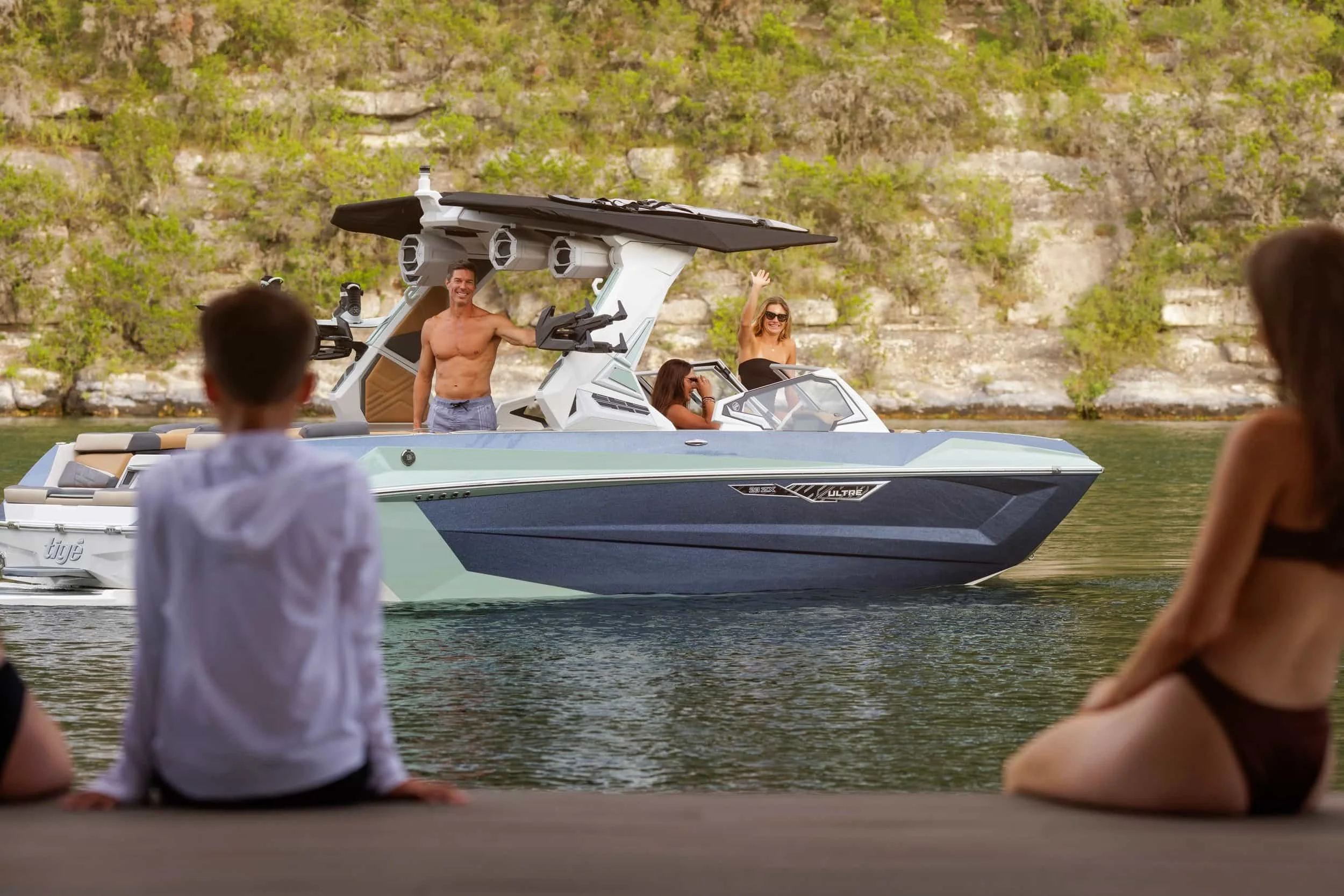 A group of people on a boat wave to two people sitting on a dock by a lake with rocky, green cliffs in the background.