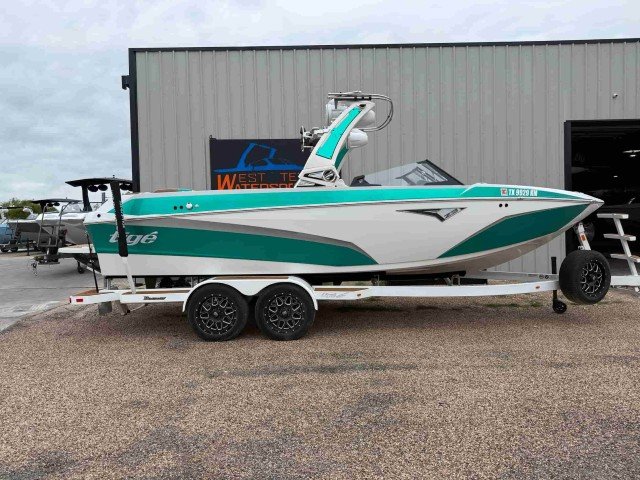 A teal and white boat on a trailer is parked outside a gray metal building with boats and a sign reading West Texas Watersports.