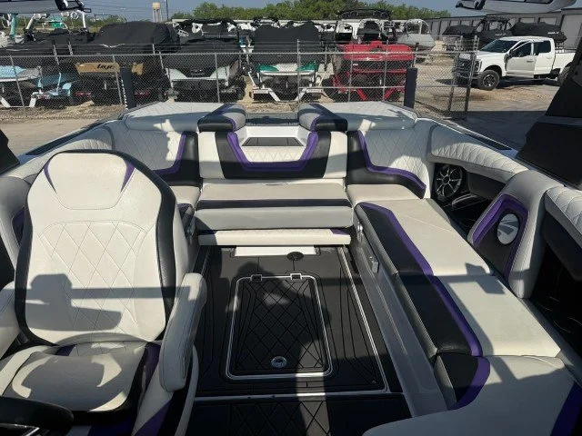 Interior of a boat with white and purple cushioned seating, a captain's chair, and a storage hatch on the deck, with outdoor vehicles and trees visible in the background.