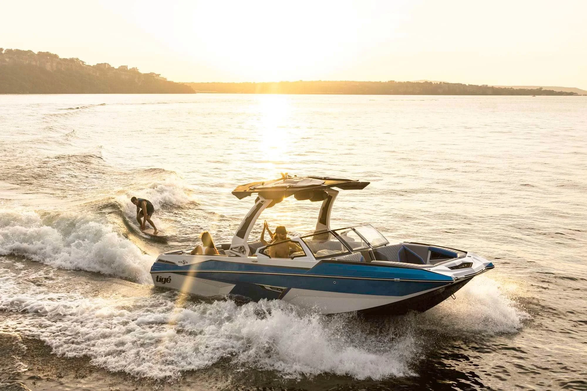 A blue and white speedboat pulls a wakeboarder across a sunlit lake with gentle waves and a distant shoreline.