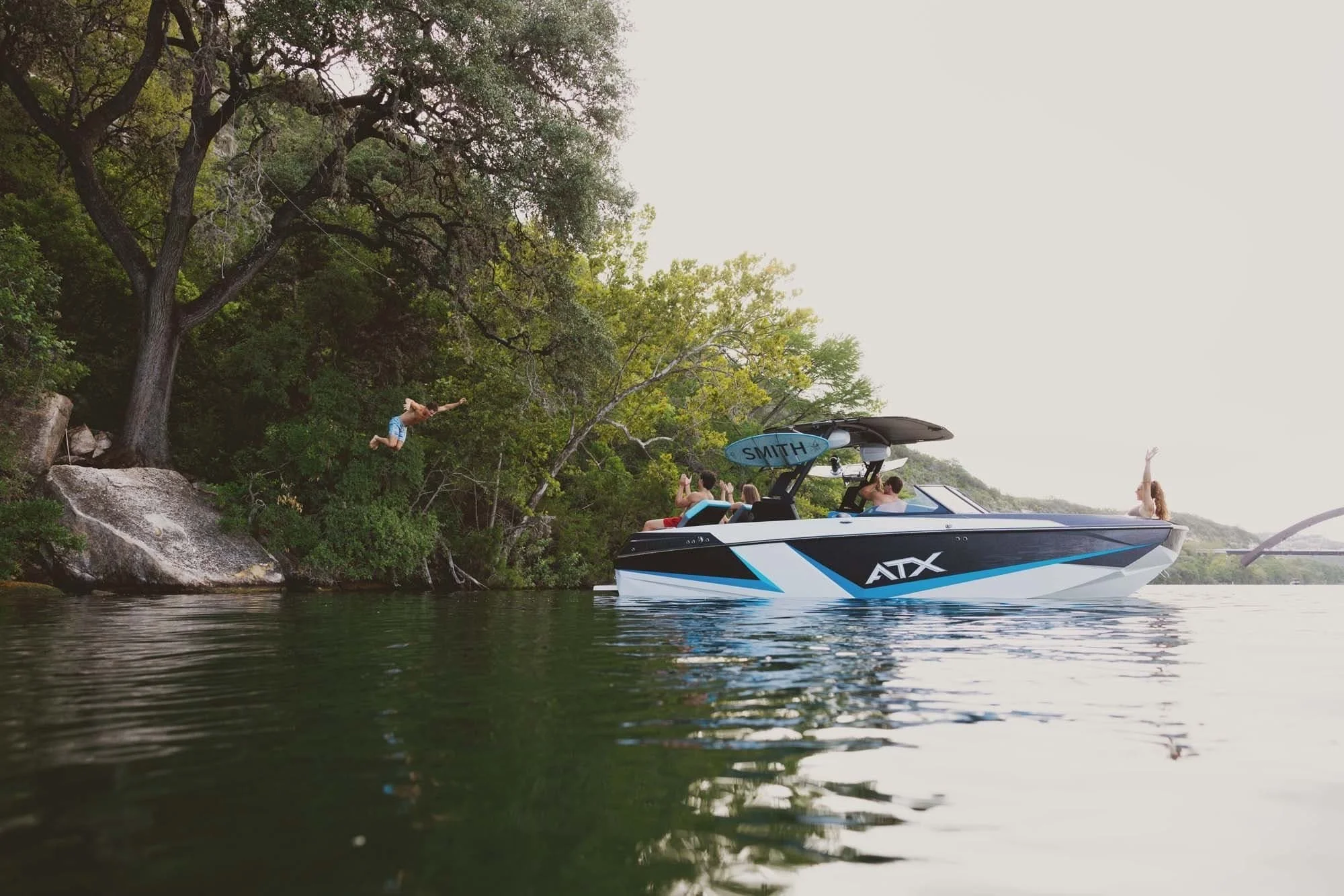 A group enjoys a day on the lake; two people jump into the water from a boat and a rock, surrounded by trees.