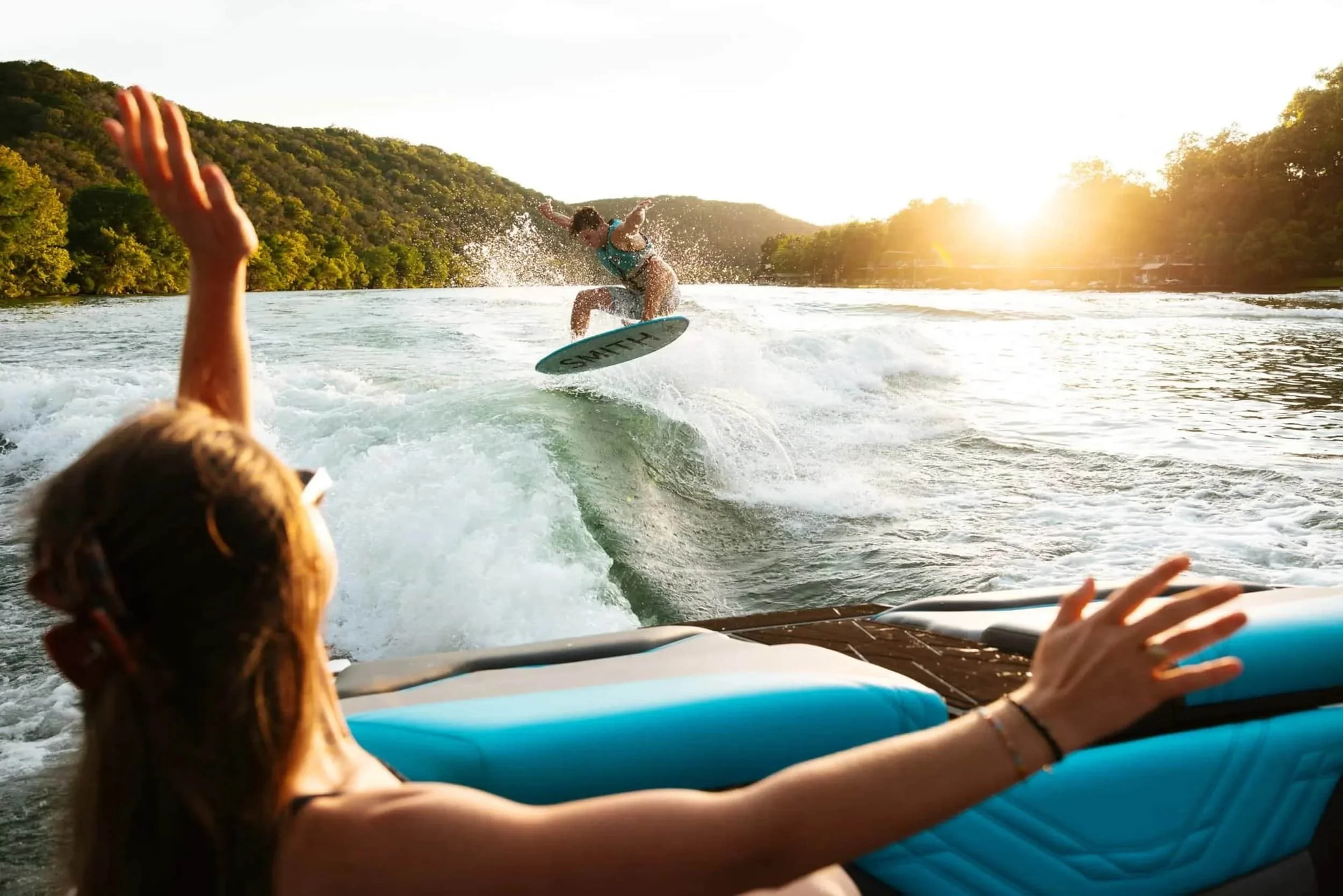 Person wakesurfing behind a boat at sunset, with another person watching and cheering from the back of the boat.