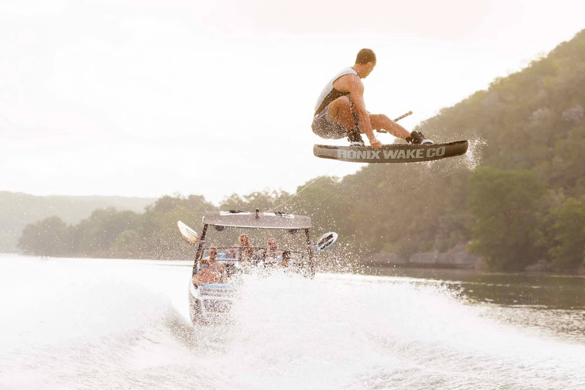 A man wakeboarding jumps high above the water, pulled by a boat with people watching and cheering on a sunny lake.