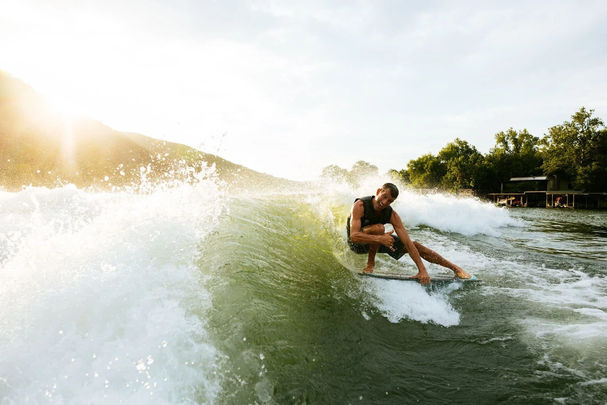 A man in a wetsuit surfs on a wave with bright sunlight and green trees in the background.