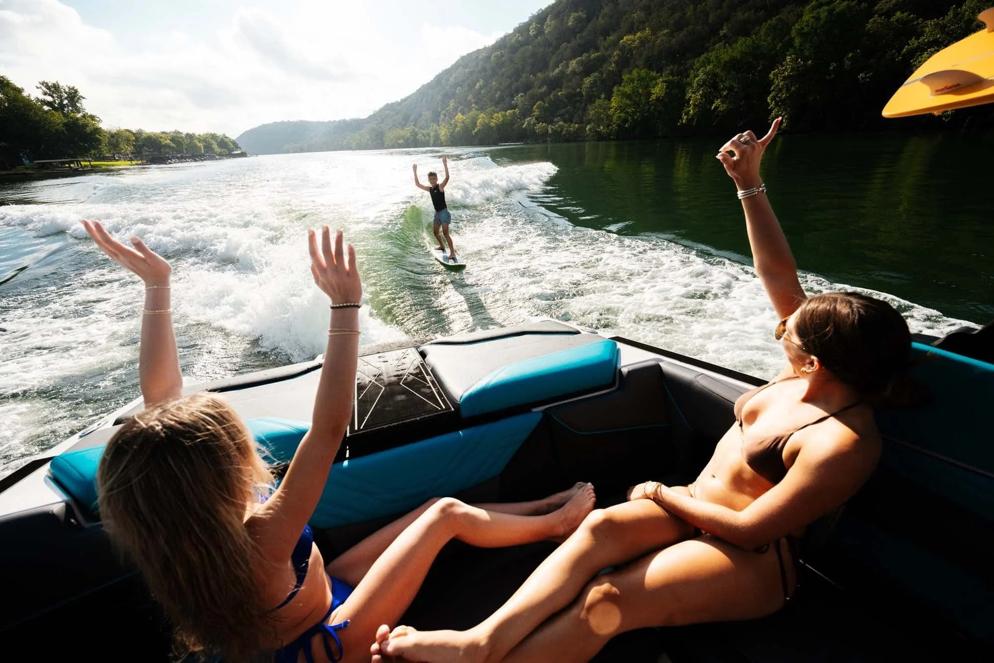 Two women on a boat cheer for a person wakeboarding behind them on a sunny day, surrounded by green hills and calm water.