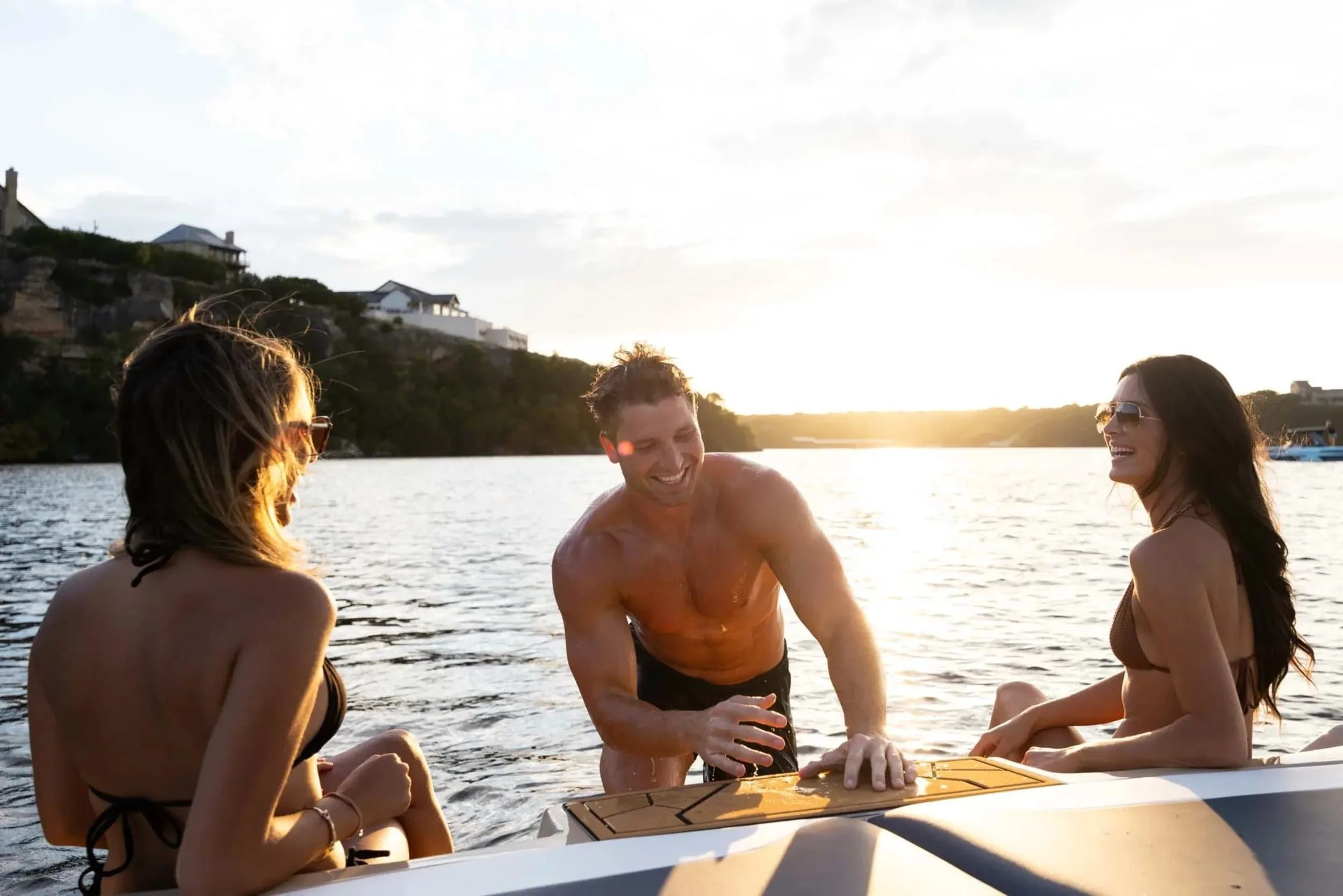 Three people in swimsuits laughing and relaxing on a boat at sunset on a lake, with houses visible in the background.