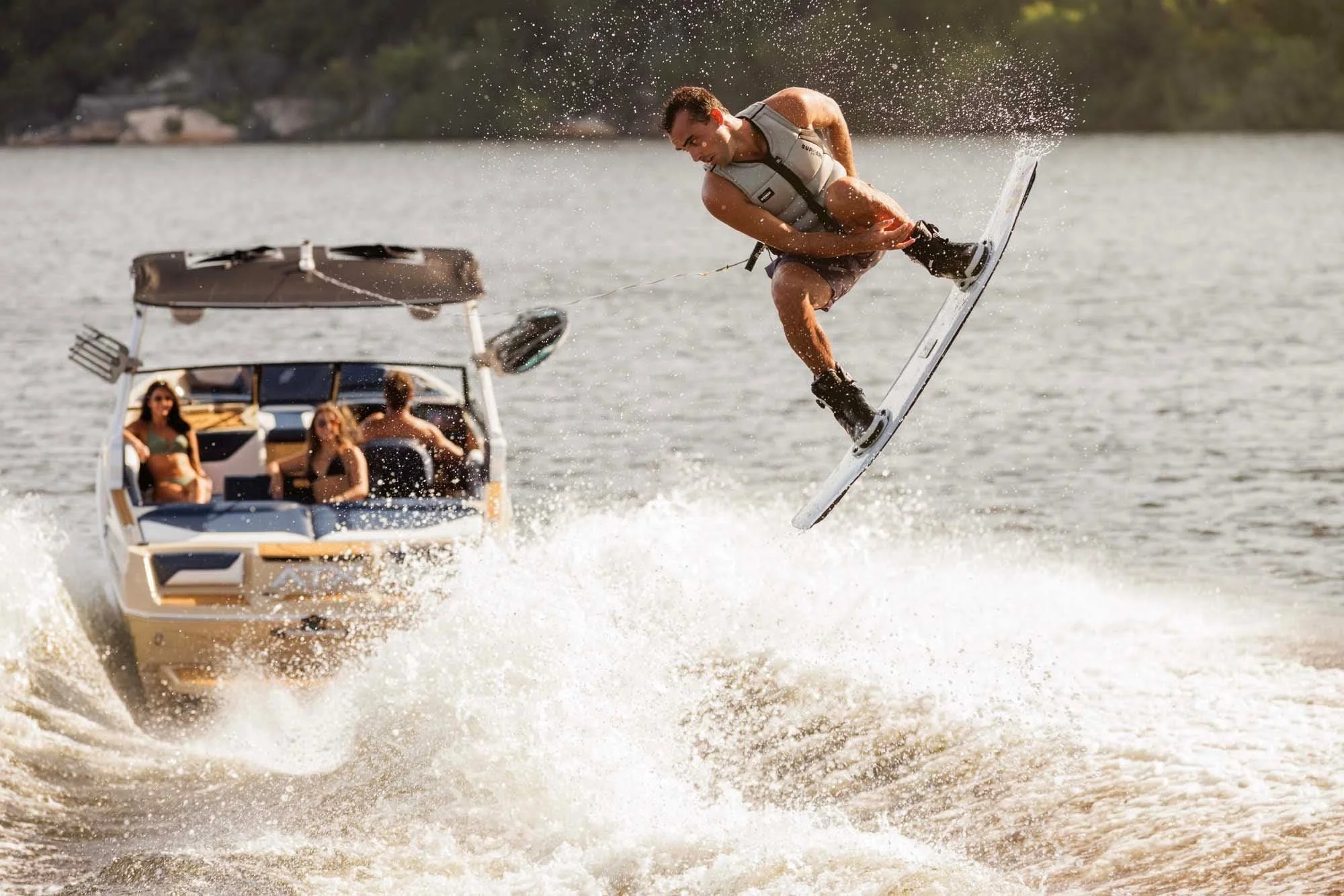 A man wakeboarding performs an aerial trick above the water, while three people watch from a boat behind him.