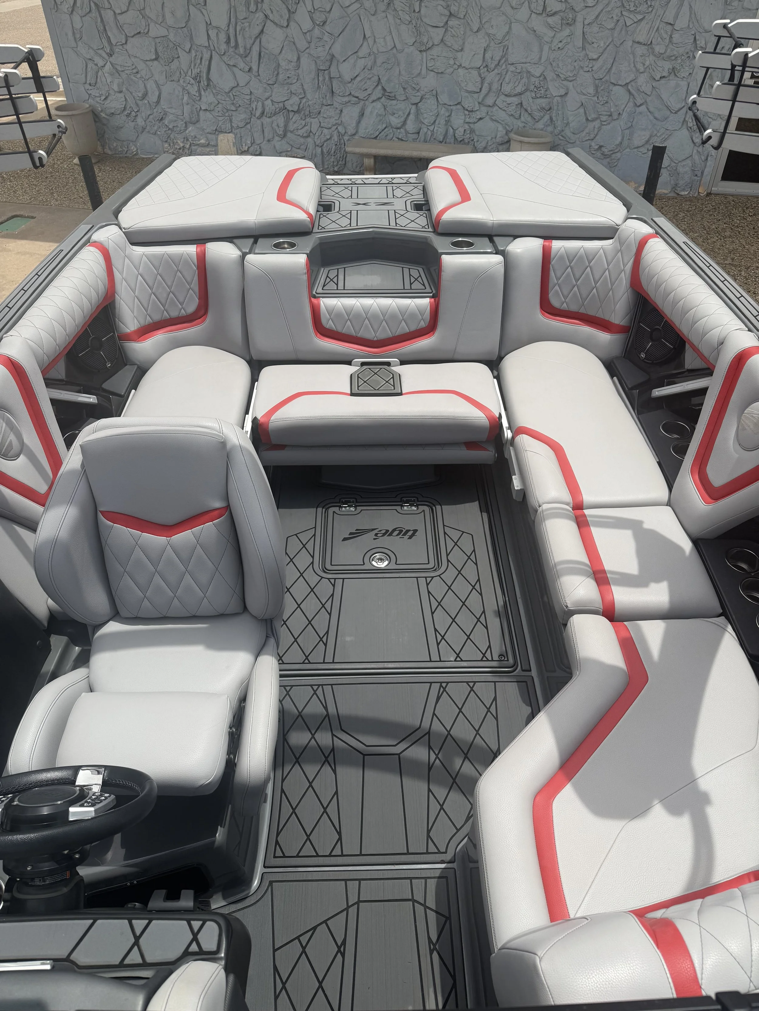 Interior of a luxury boat with white and red cushioned seating, a black steering wheel, and gray flooring with geometric patterns.