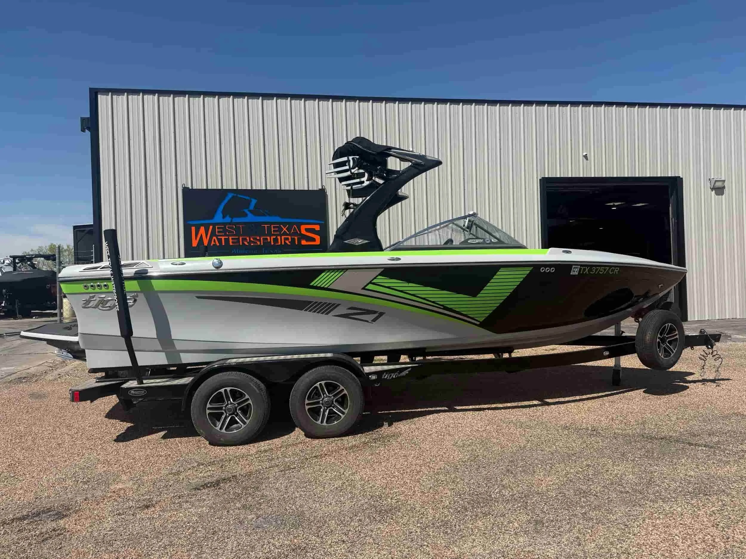 A black, green, and white speedboat on a trailer is parked outside a West Texas Watersports building on a sunny day.