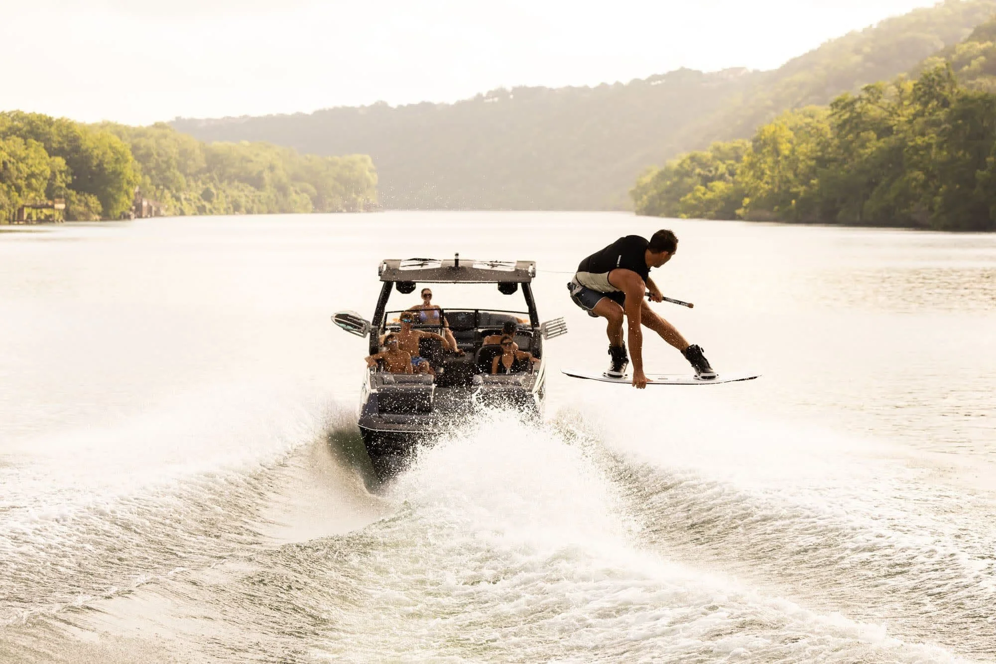 A person wakeboarding and doing a jump behind a boat with people watching, on a lake surrounded by trees.