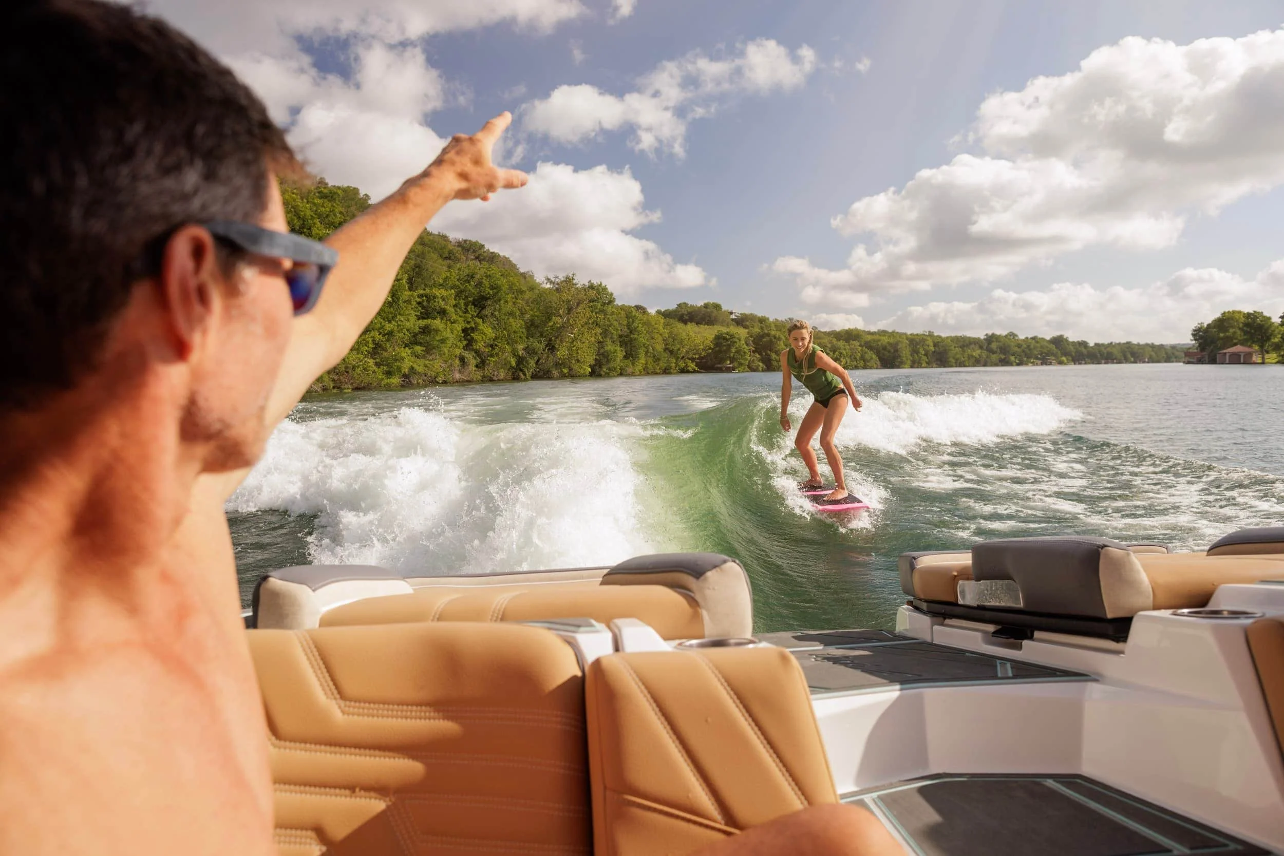 A man on a boat points toward a woman wakesurfing behind the boat on a sunny day with trees in the background.