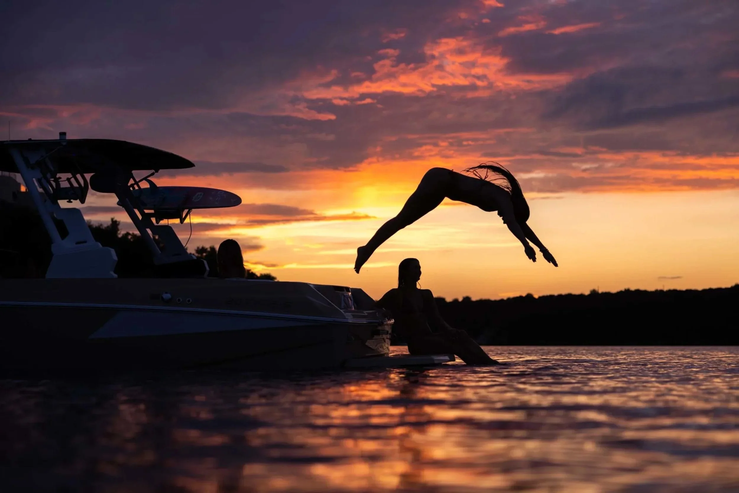 Person dives off a boat into a lake at sunset, with vibrant orange and purple clouds in the sky.