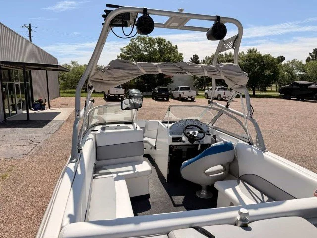 The picture shows the interior and dashboard of a white motorboat with a canopy, seating, steering wheel, and speakers, parked outdoors on a gravel lot with trees and a building in the background.