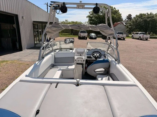 White motorboat with gray seating, steering wheel, and wakeboard tower, parked in a lot with a building and several cars in the background.