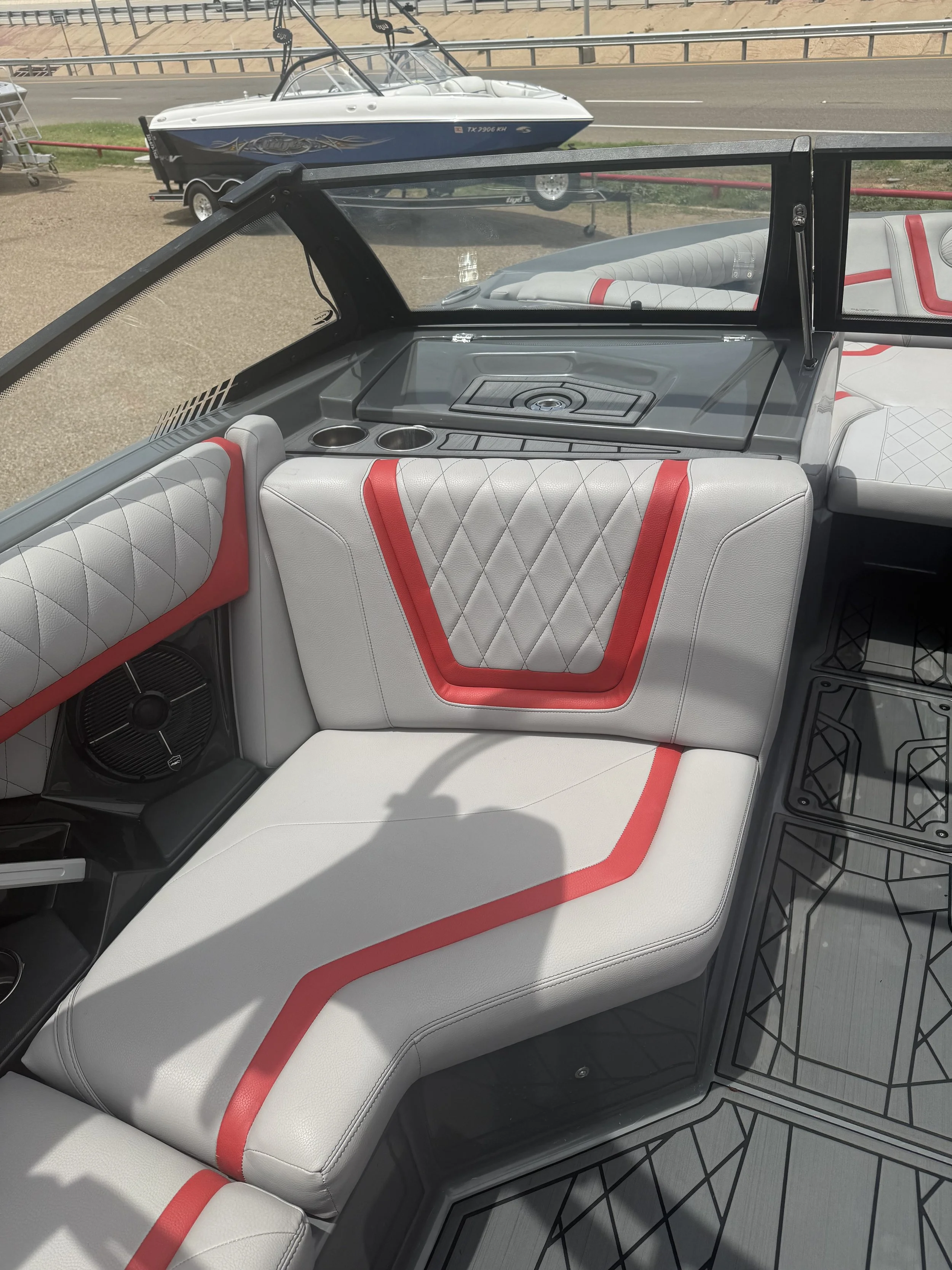 The image shows the interior of a boat with white and red cushioned seats and a grey dashboard. Through the windshield, a blue and white wakeboarding boat on a trailer is visible parked on a dirt area near a road, with a guardrail and some sandy terr