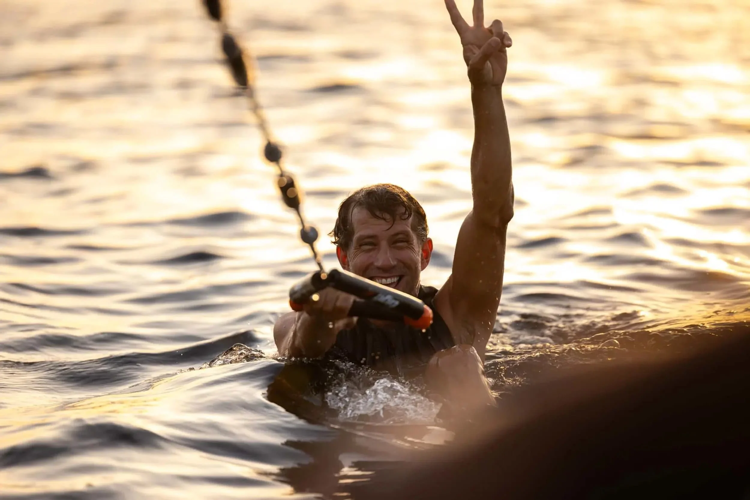 Man in water holding a tow handle and smiling, raising one hand with a peace sign at sunset.