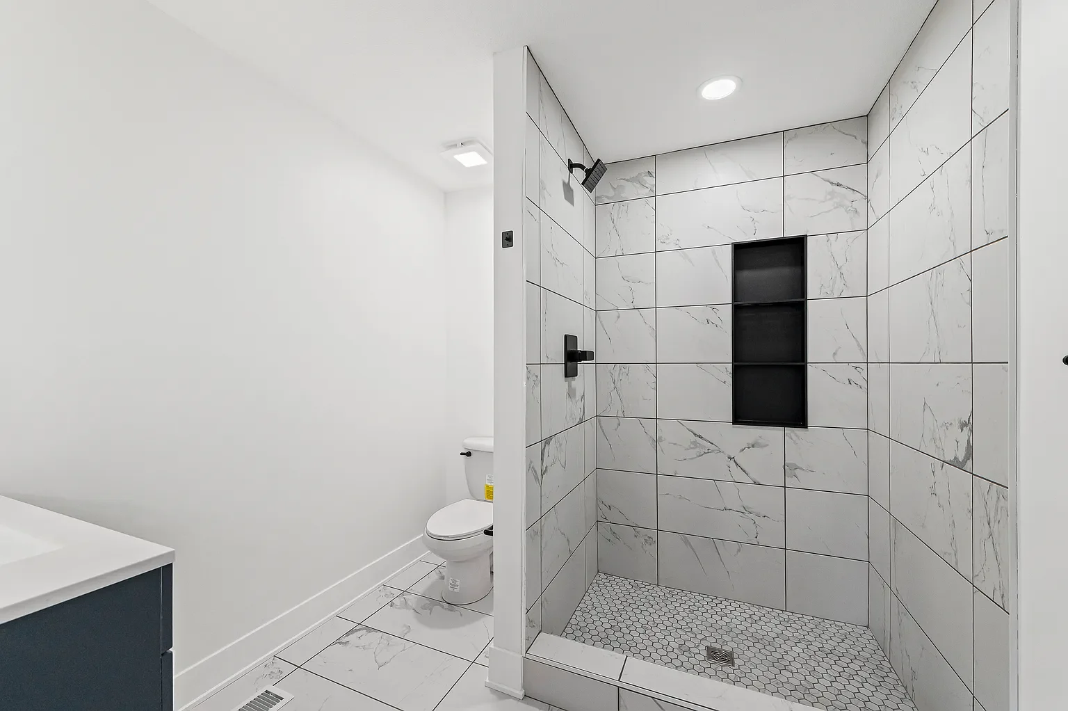 Modern bathroom with white marble tile in the shower area, featuring a black showerhead, black controls, a built-in black shelf, and a white toilet with a closed lid.