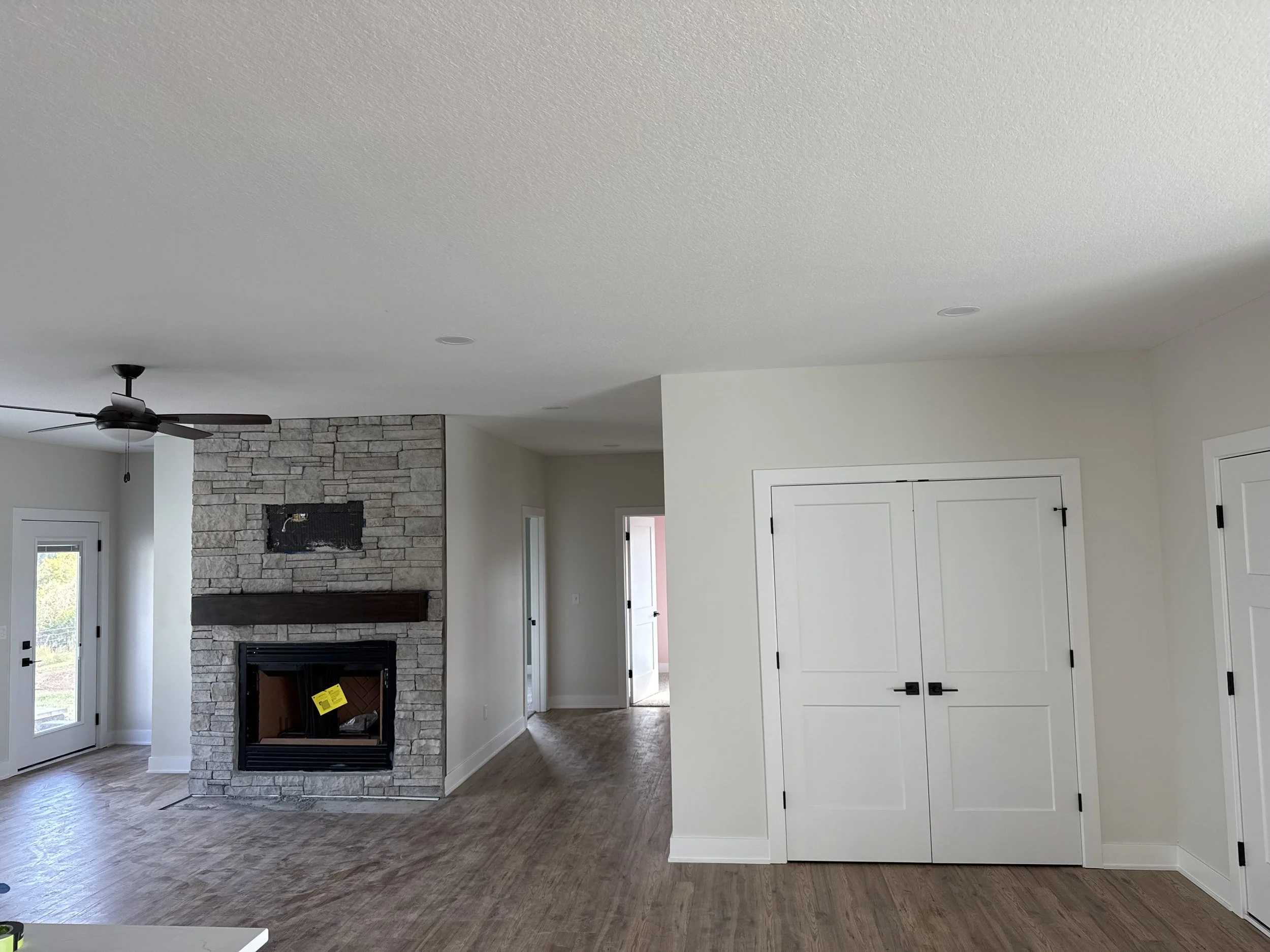 Empty living room with a stone fireplace, ceiling fan, white walls, hardwood flooring, and white double doors.