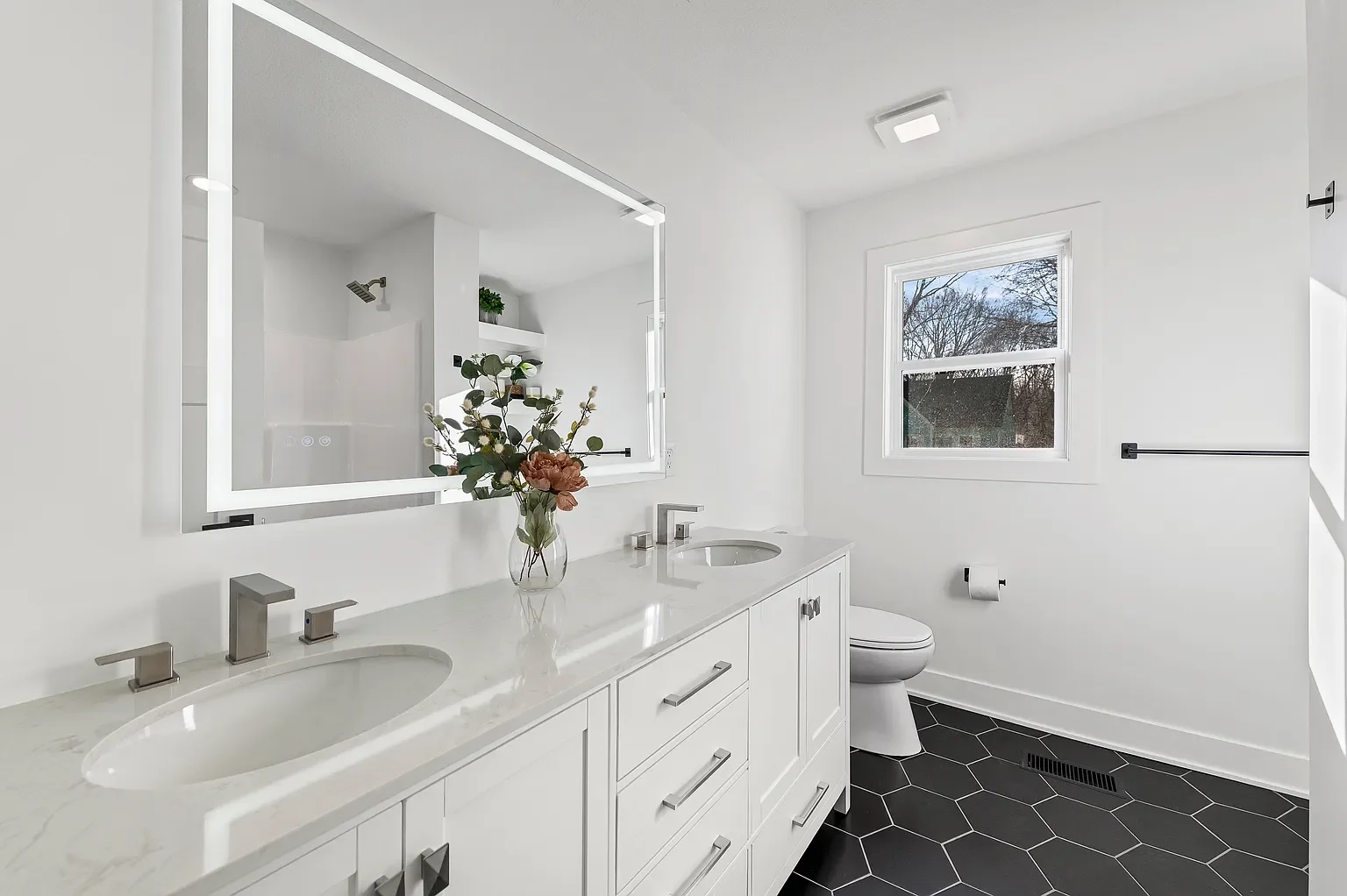 Modern white bathroom with double sink vanity, large mirror with LED lighting, black hexagon floor tiles, and a window showing trees outside.
