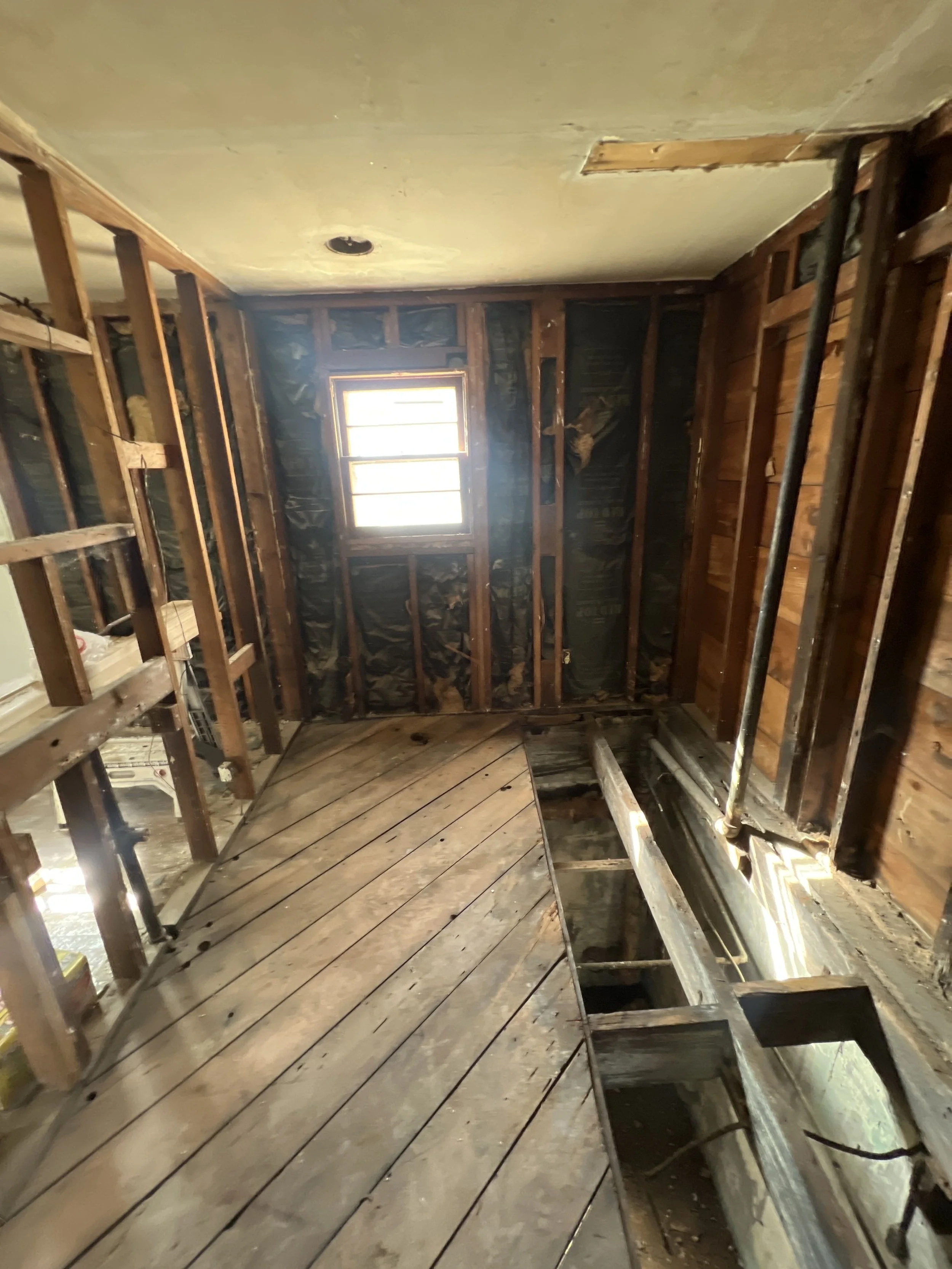 Room under renovation with exposed wooden framing, a small window, unfinished flooring, and visible pipes and wires.