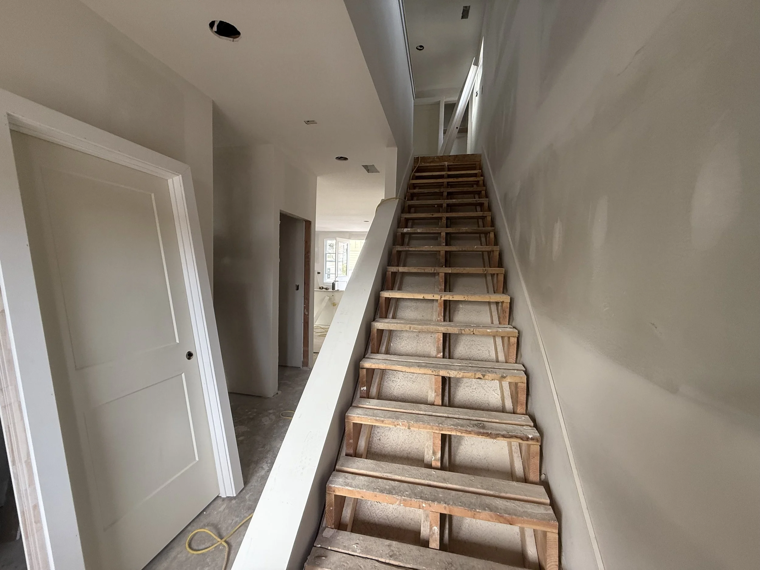 Interior of a house under construction with an unfinished staircase made of wooden frame, adjacent walls partially painted, and an open ceiling with exposed hole