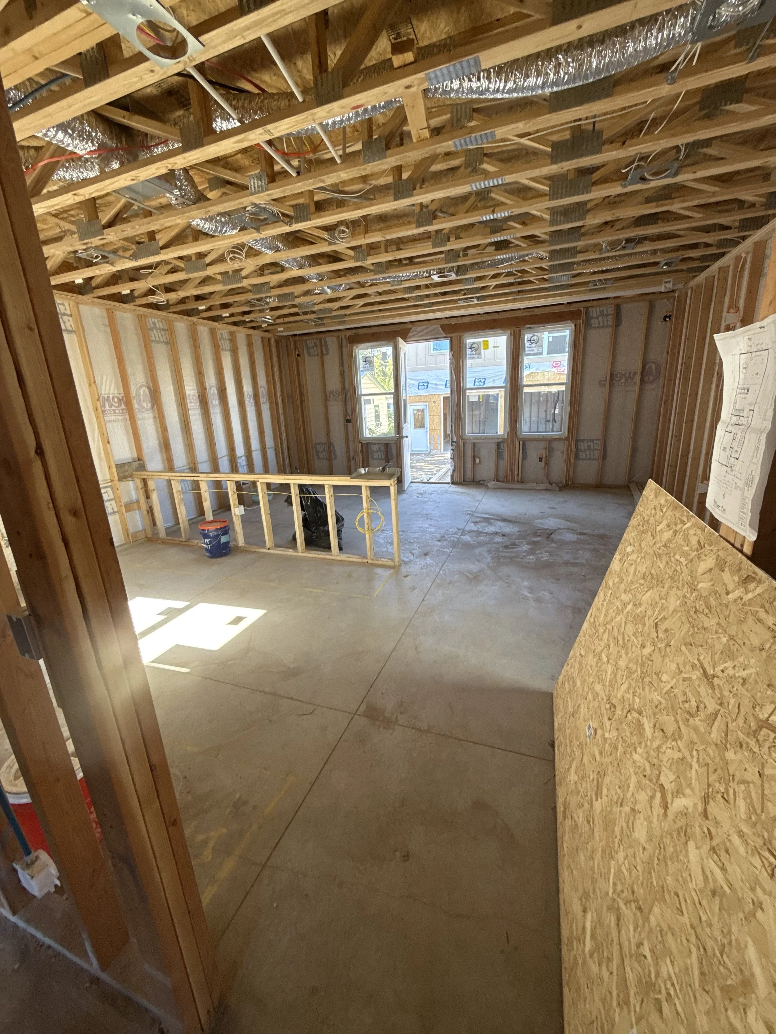 Interior of a house under construction with wooden framing, exposed ceiling with ducts and wires, and large windows at the far end.