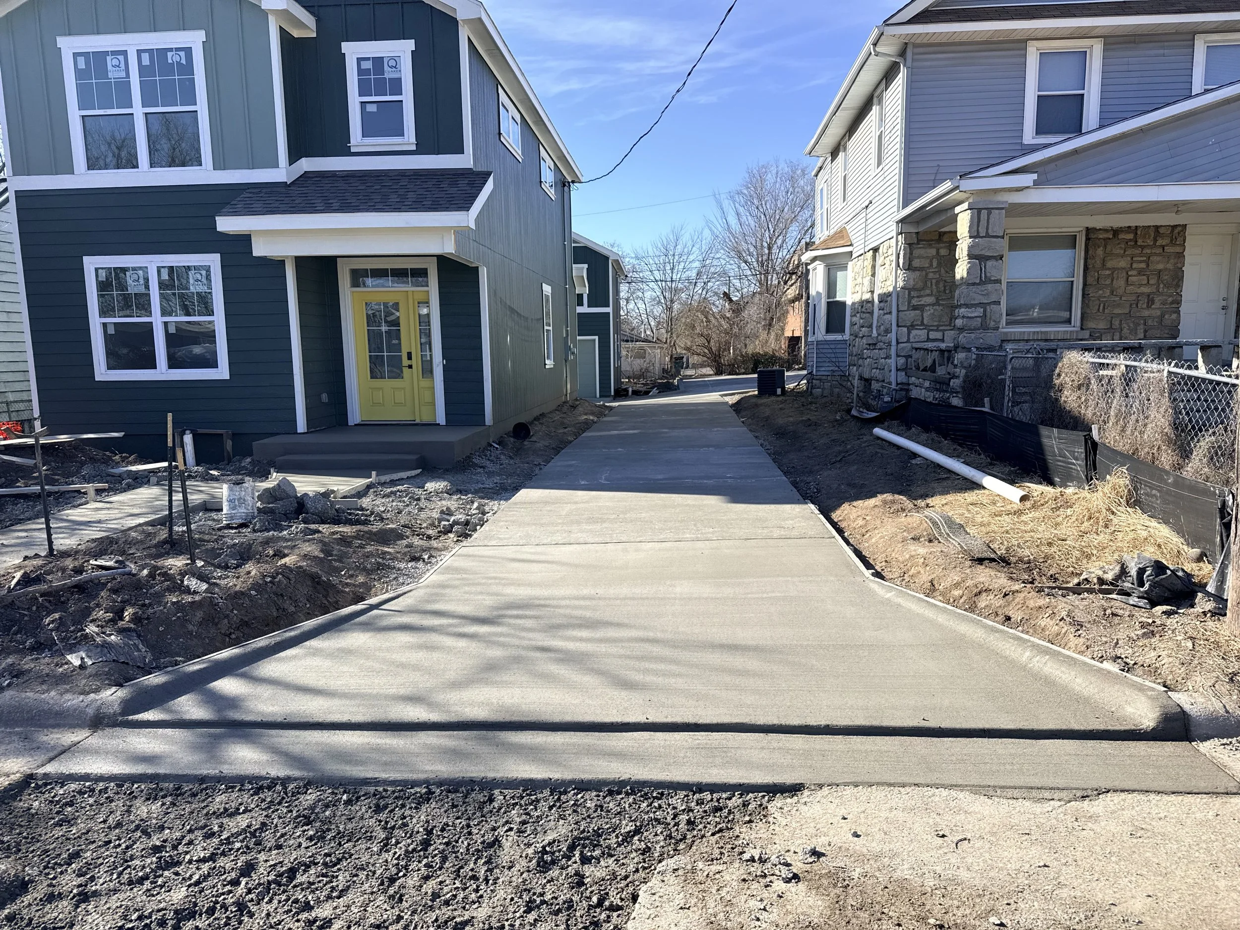 Newly poured concrete sidewalk between two residential houses, with construction materials and dirt on the sides, in a neighborhood with clear skies.