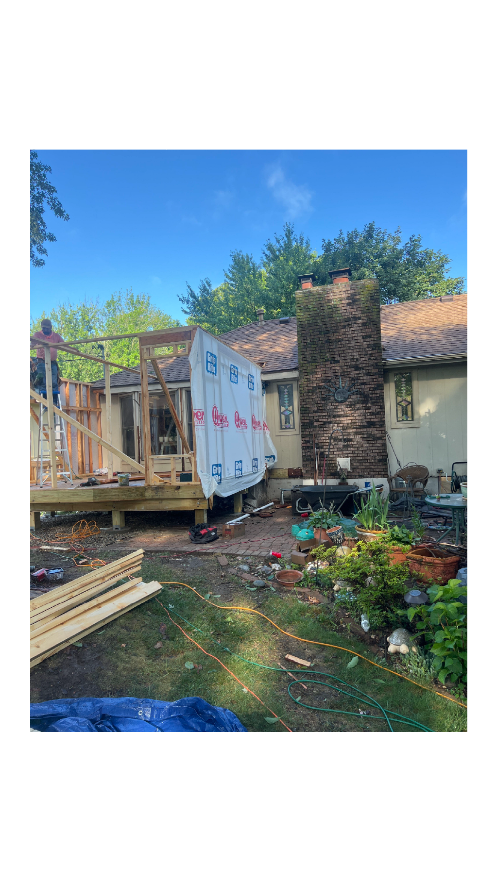 A house undergoing construction with a new wooden deck being built on the back, tools and construction materials scattered around, and a person working on the deck. The backyard has plants and garden decorations next to the house.