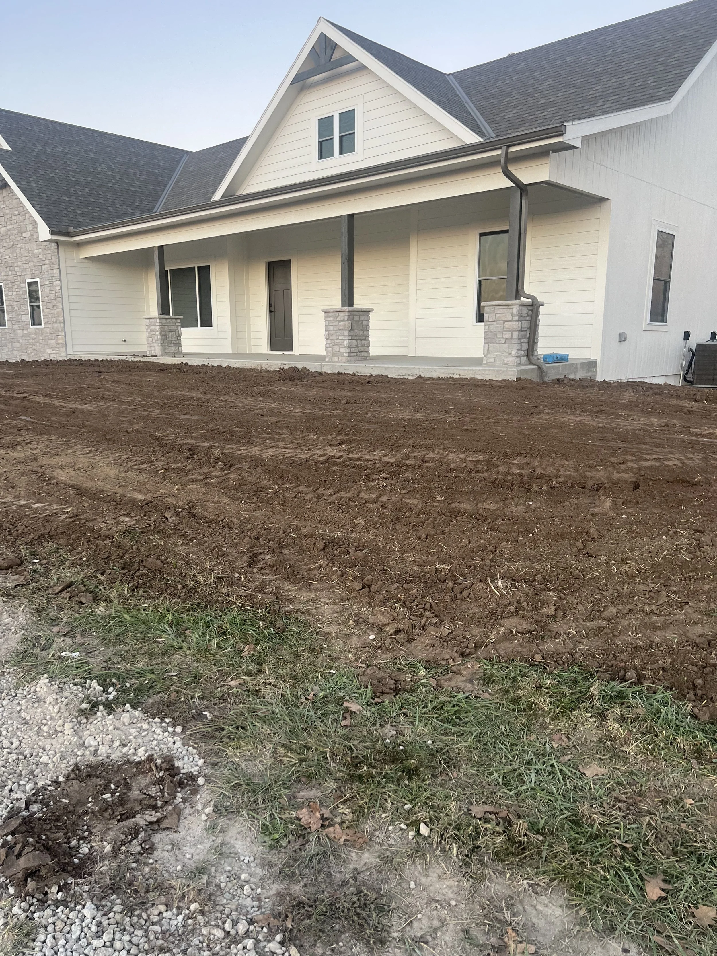 Newly built white house with a porch, gray roof, and stone pillars, surrounded by bare soil and grass.
