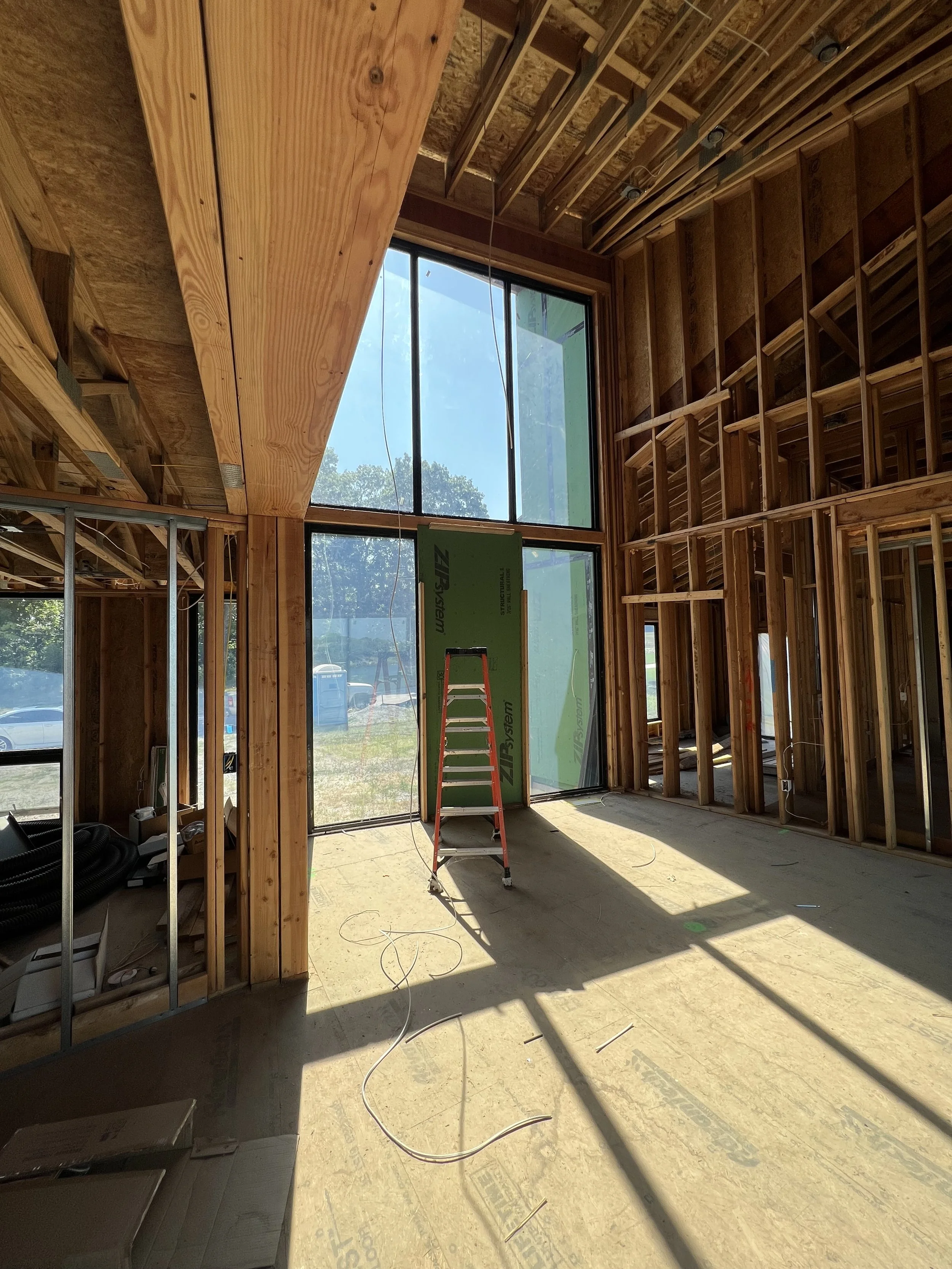 Interior of a house under construction with exposed wooden framing, a large window showing trees outside, a red ladder, and construction materials scattered on the floor.