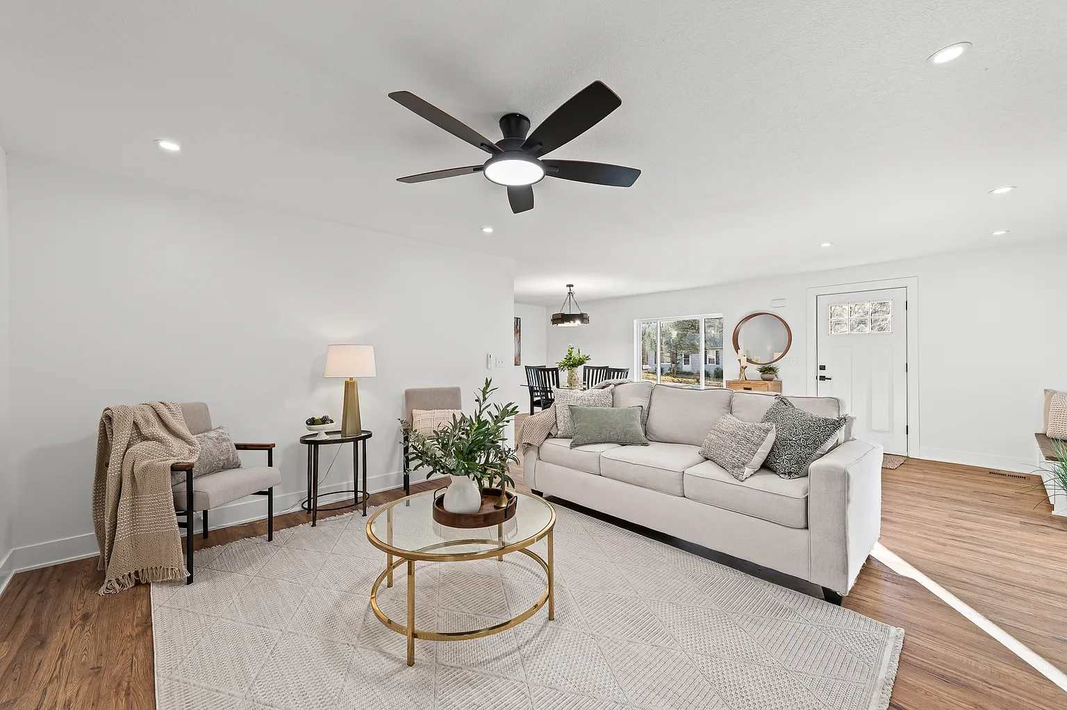 Bright living room with white walls, hardwood floors, a white sofa with pillows, a round coffee table with a plant, a beige armchair, a side table with a lamp, and a dining area in the background.