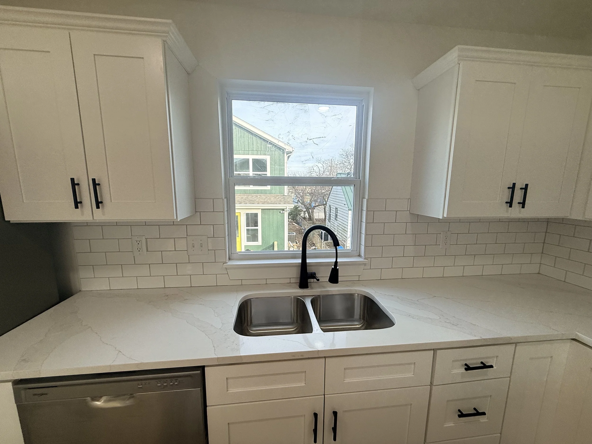 Kitchen with white cabinets, white marble countertop, stainless steel sink, black faucet, white subway tile backsplash, window above the sink, and outside view of houses.