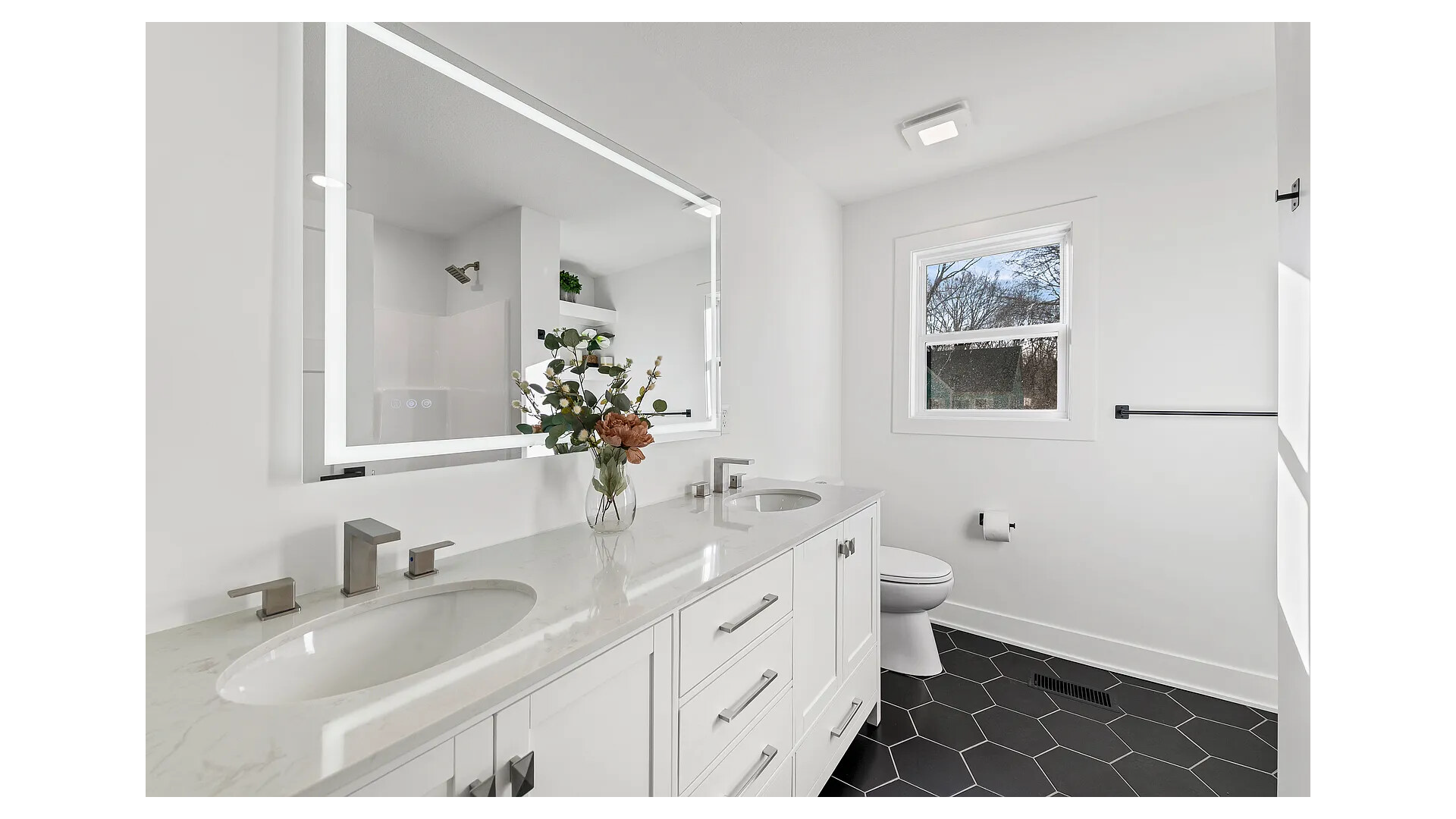 Modern white bathroom with double vanity, black hexagonal floor tiles, and a large mirror with built-in lighting.