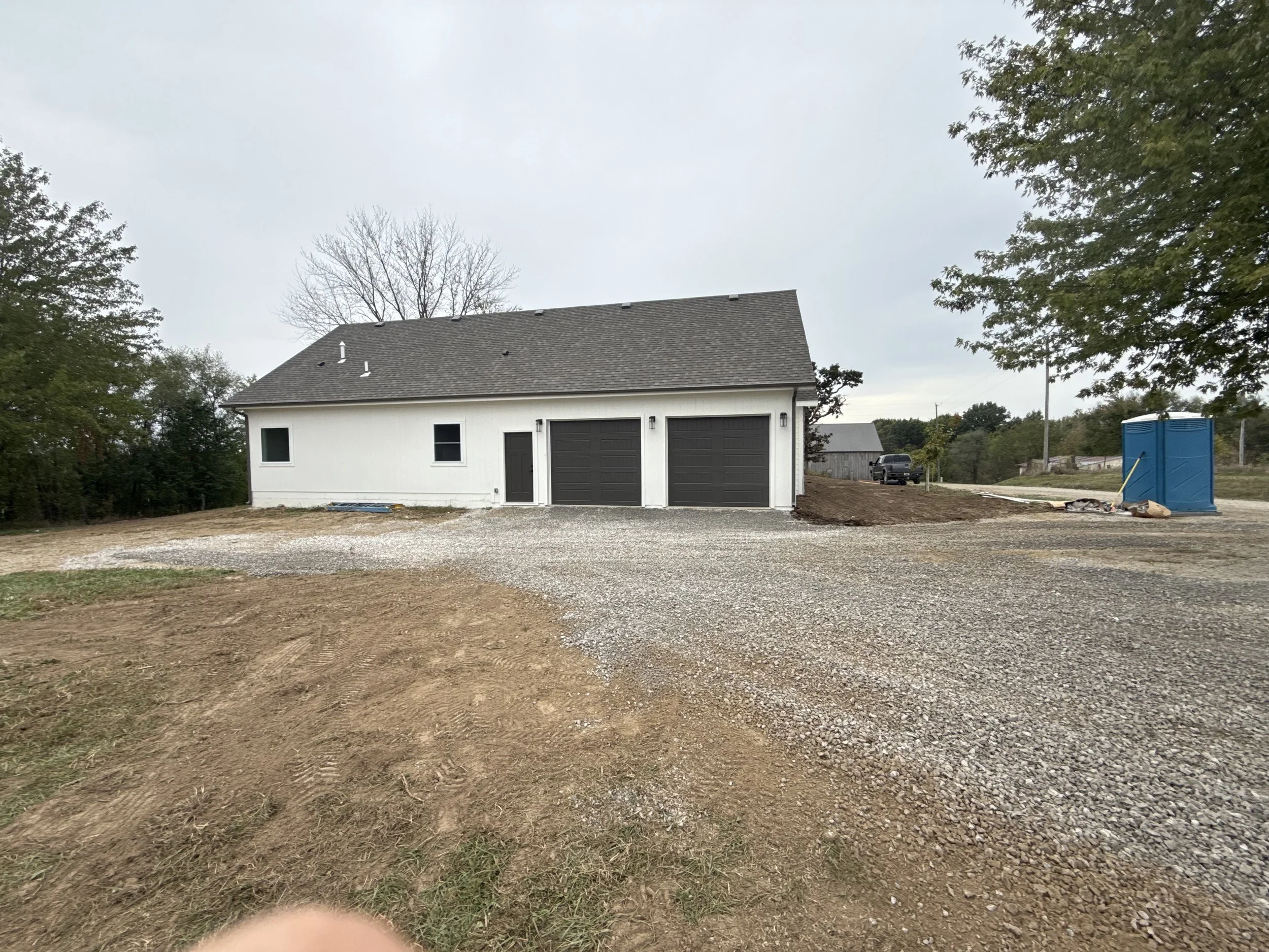 Newly constructed house with a white exterior, black garage doors, and a gravel driveway, surrounded by a yard with dirt patches, trees, and a portable toilet on the right.