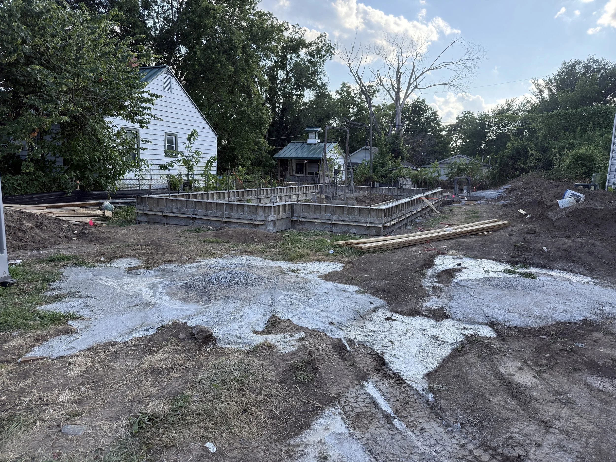 Construction site with framing for a new foundation, surrounded by dirt and gravel, in a residential area with houses and trees in the background.