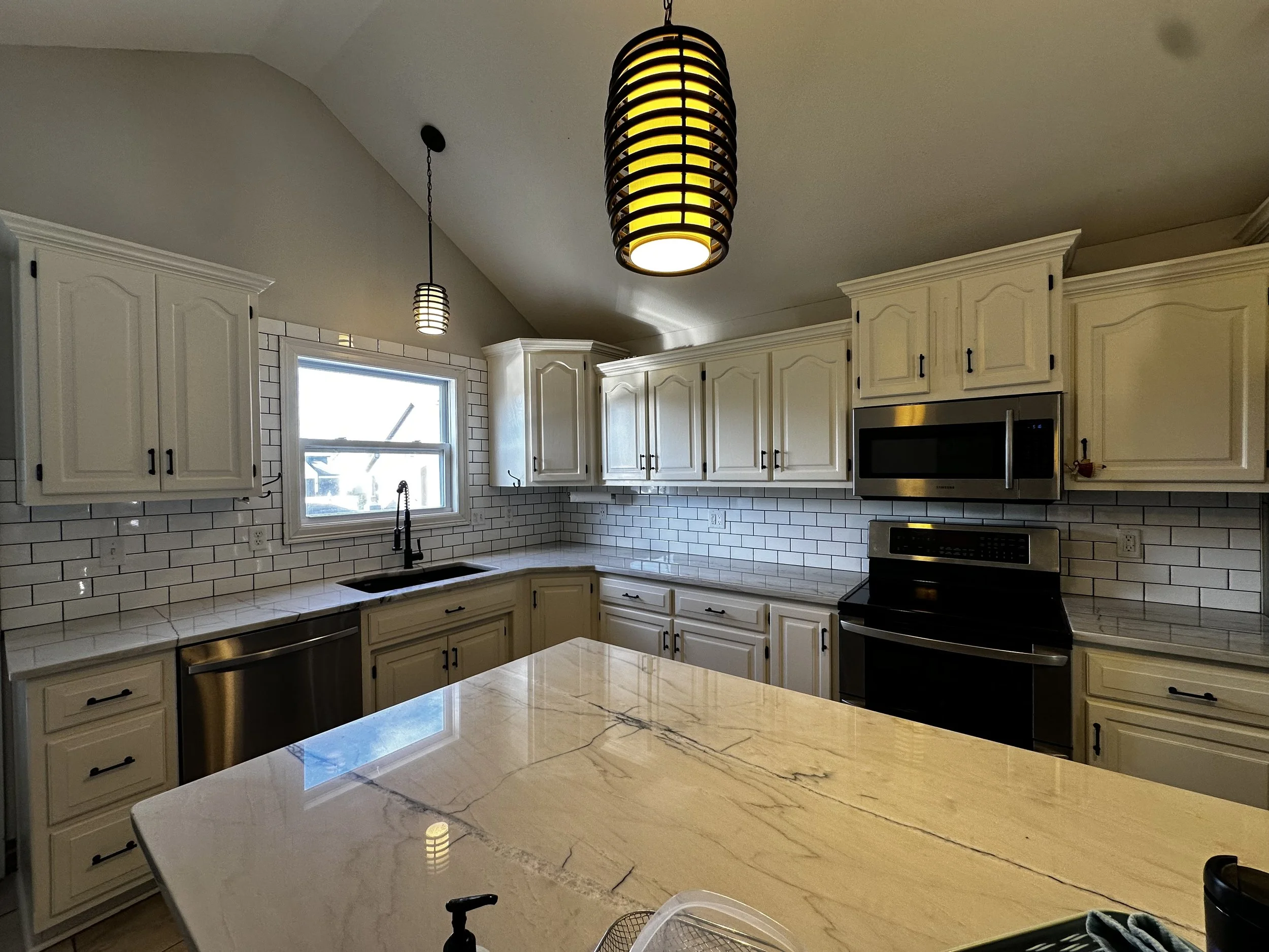 A modern kitchen with white cabinets, subway tile backsplash, a marble countertop, and stainless steel appliances including a microwave and oven. There are two pendant lights hanging from the ceiling and a window over the sink.