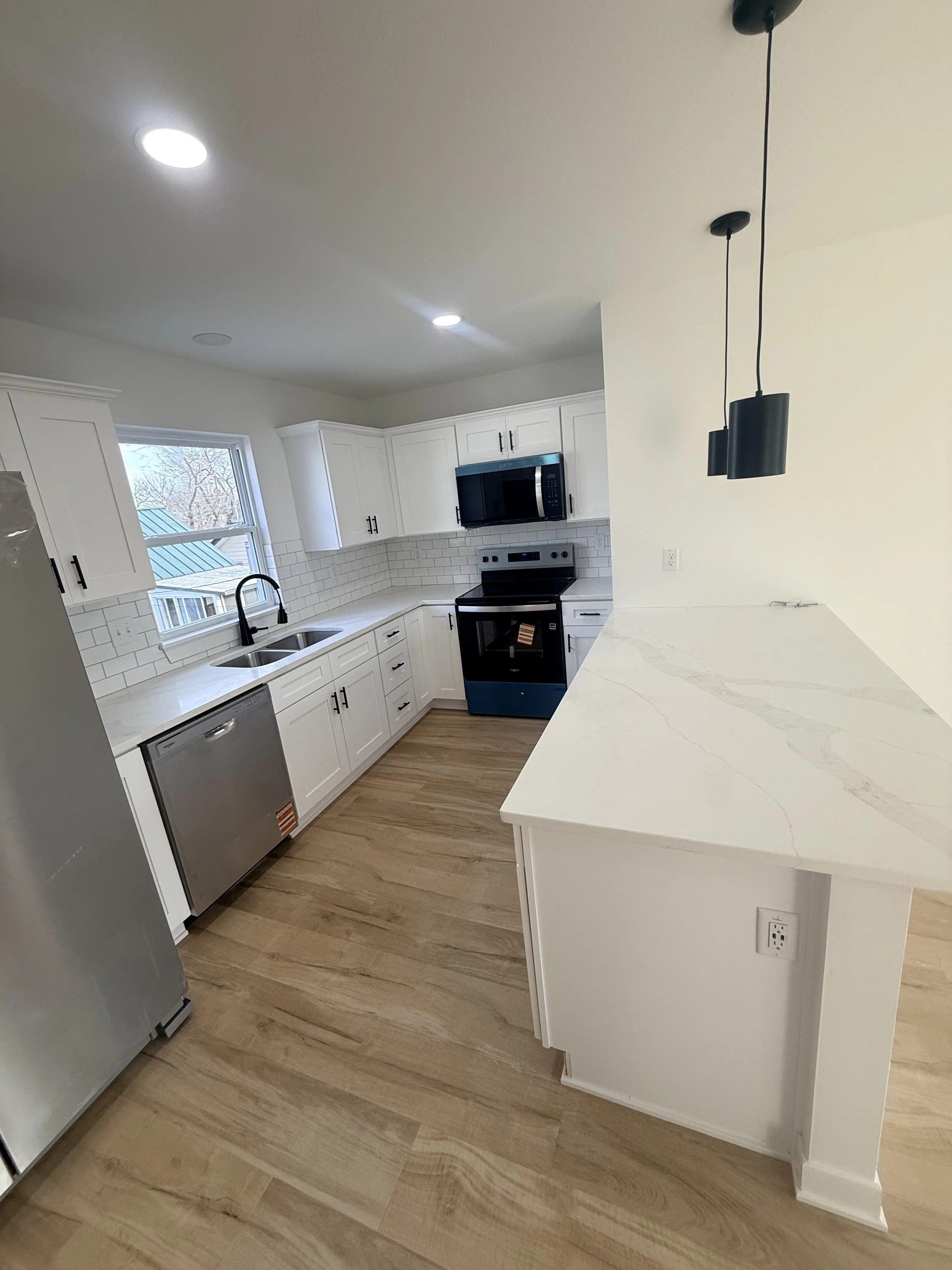 Modern kitchen with white cabinets, stainless steel appliances, a window above the sink, and a light-colored countertop with a marble pattern. Wooden flooring and black pendant lights hanging over a breakfast bar.