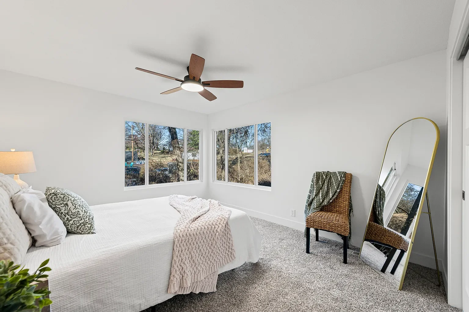Bright bedroom with white walls, a large bed with white linens and decorative pillows, a nightstand with a lamp, a wicker chair with a throw blanket, a large mirror, and windows showing trees outside.